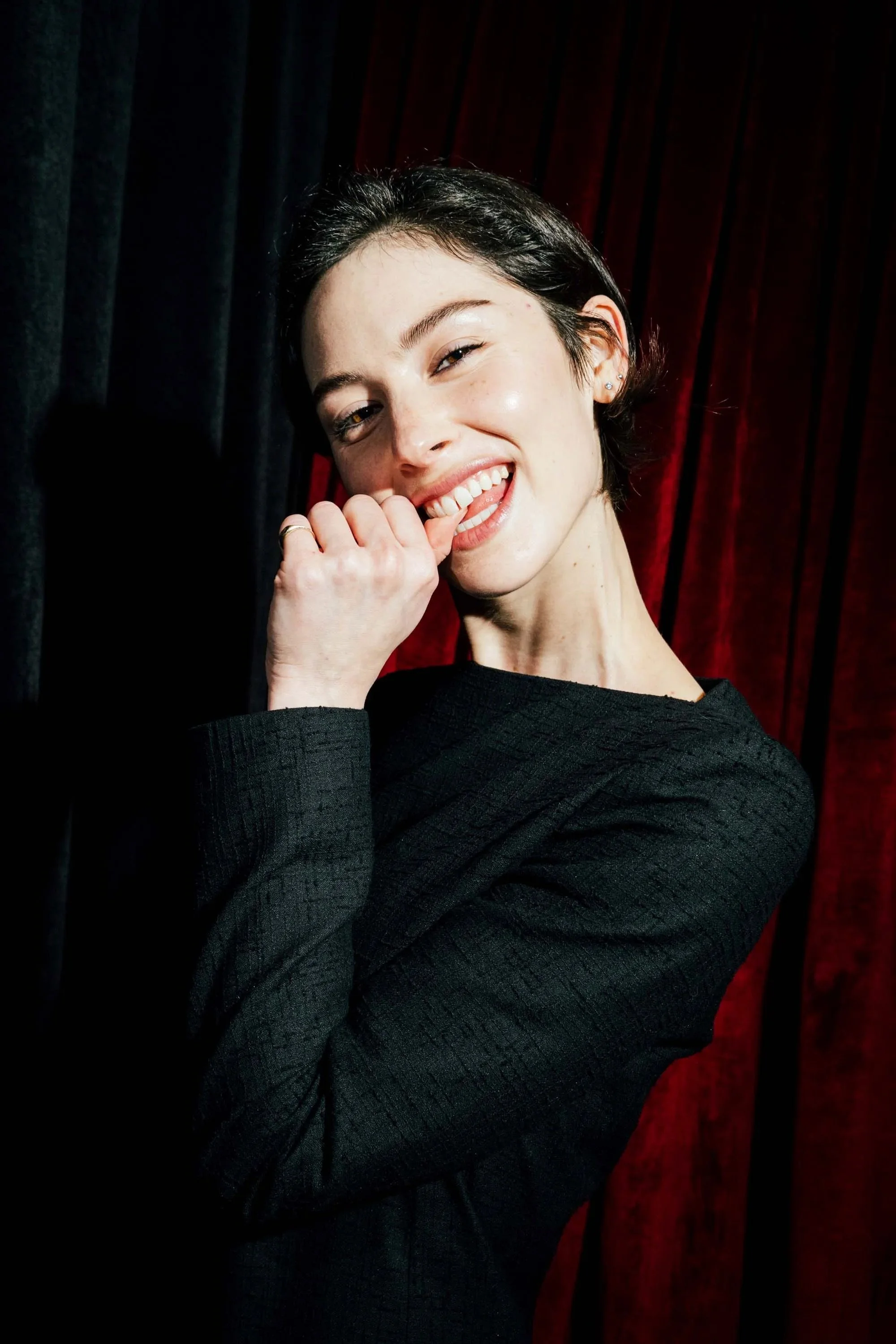 A woman with short dark hair, smiling and holding her finger near her lips, wearing a black textured shirt, against a black and red curtain background.