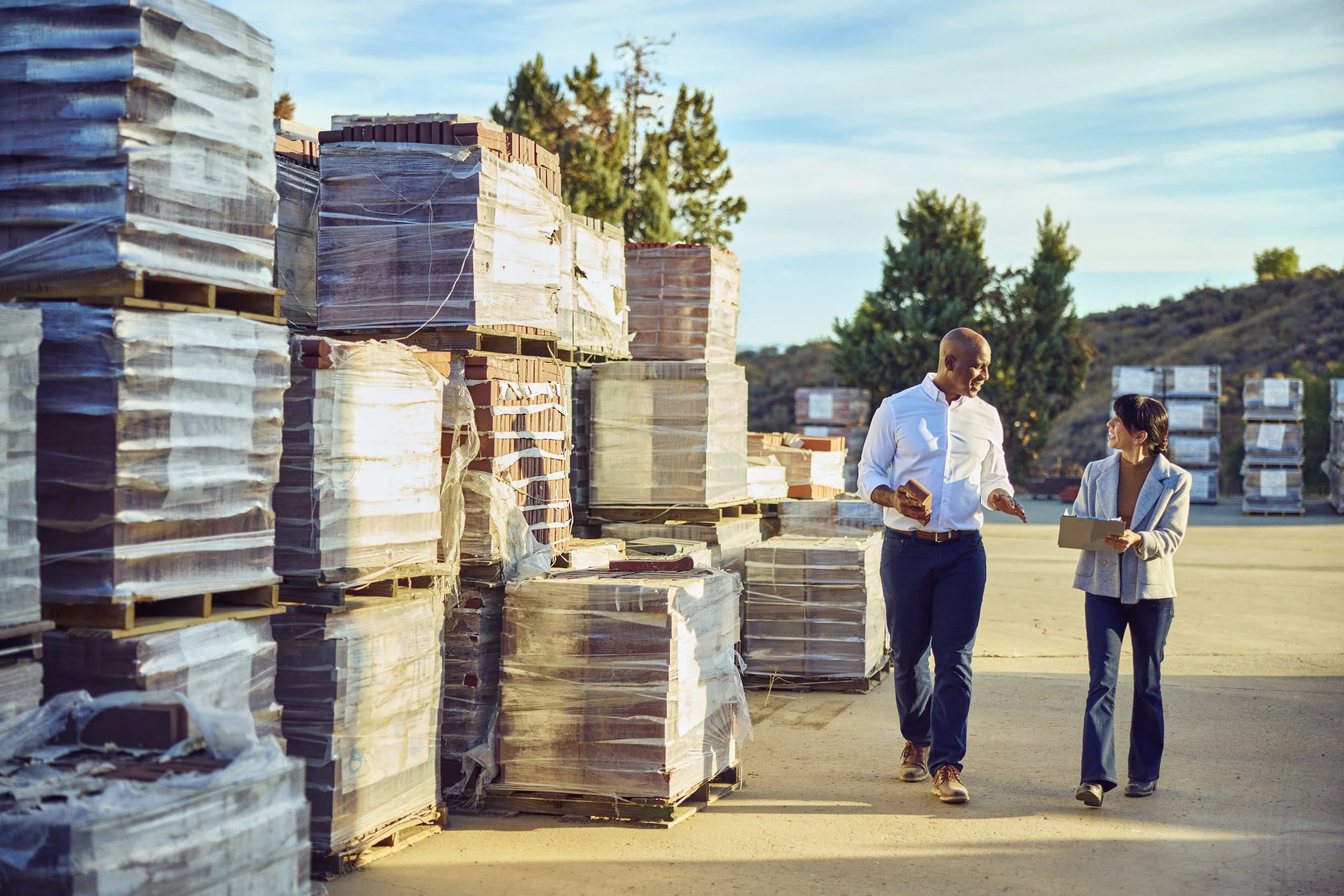 Two people, a man and a woman, walking and talking in an outdoor storage yard with stacks of wrapped building materials on pallets, trees, and a blue sky in the background.