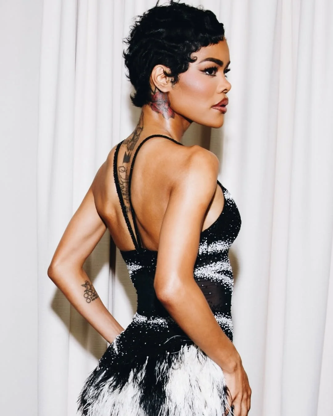 Side profile of woman with short black curly hair, tattoos on neck and arm, wearing a black and white dress with feathers, standing against white curtains.