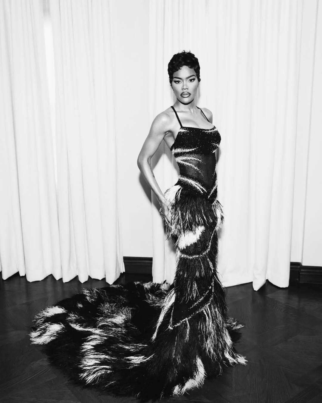 A woman in a glamorous, black and white feathered evening gown with sequins, standing in front of closed white curtains on a wooden floor.