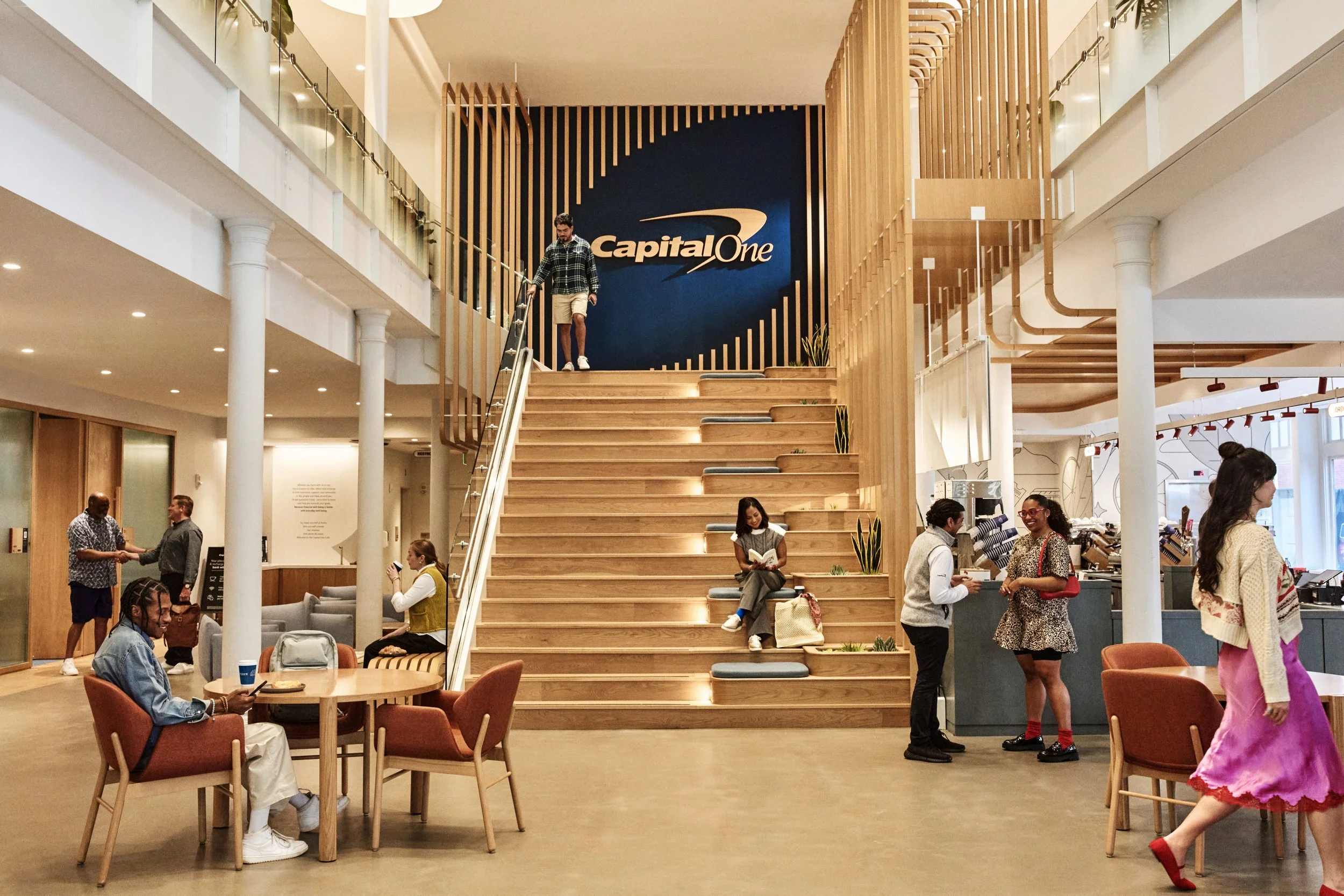 Interior of a modern, multi-story building with people sitting, standing, and walking. A staircase with wooden steps and cushions leads to a second level with a large blue wall featuring the 'Capital One' logo. People are engaged in conversations, reading, and using their phones.