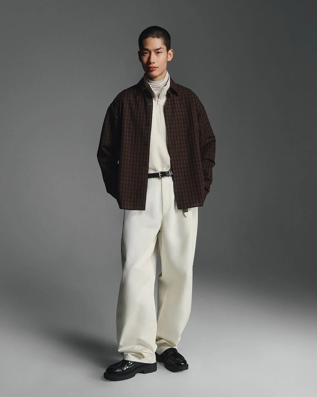 A young man with short dark hair poses against a gray background, wearing a dark plaid button-up shirt over a white zip-up top, with white wide-leg trousers and black chunky shoes.