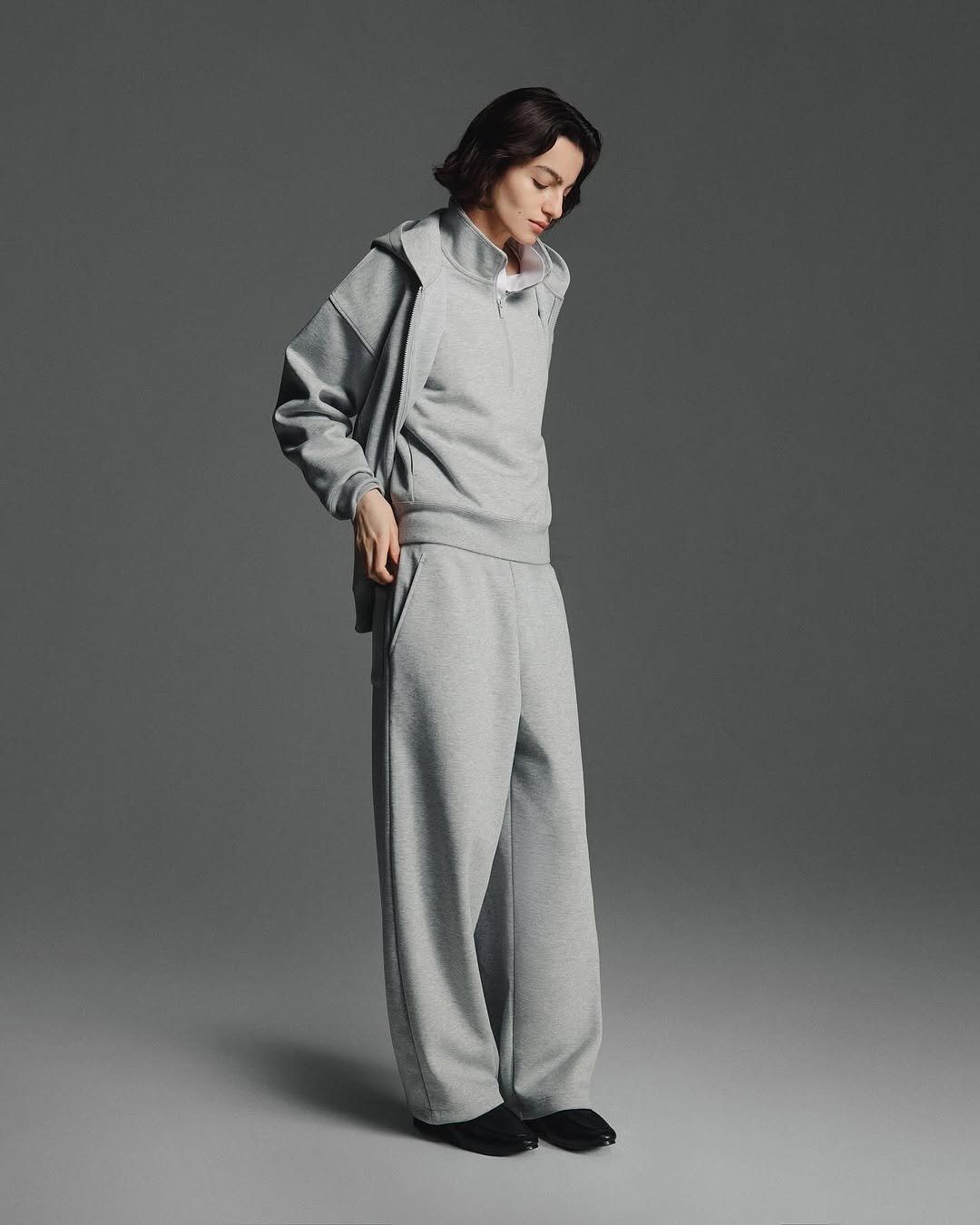 A woman standing against a gray background, dressed in a light gray tracksuit with a zip-up hoodie, matching sweatpants, and black shoes, with her head slightly bowed and hands in her pockets.