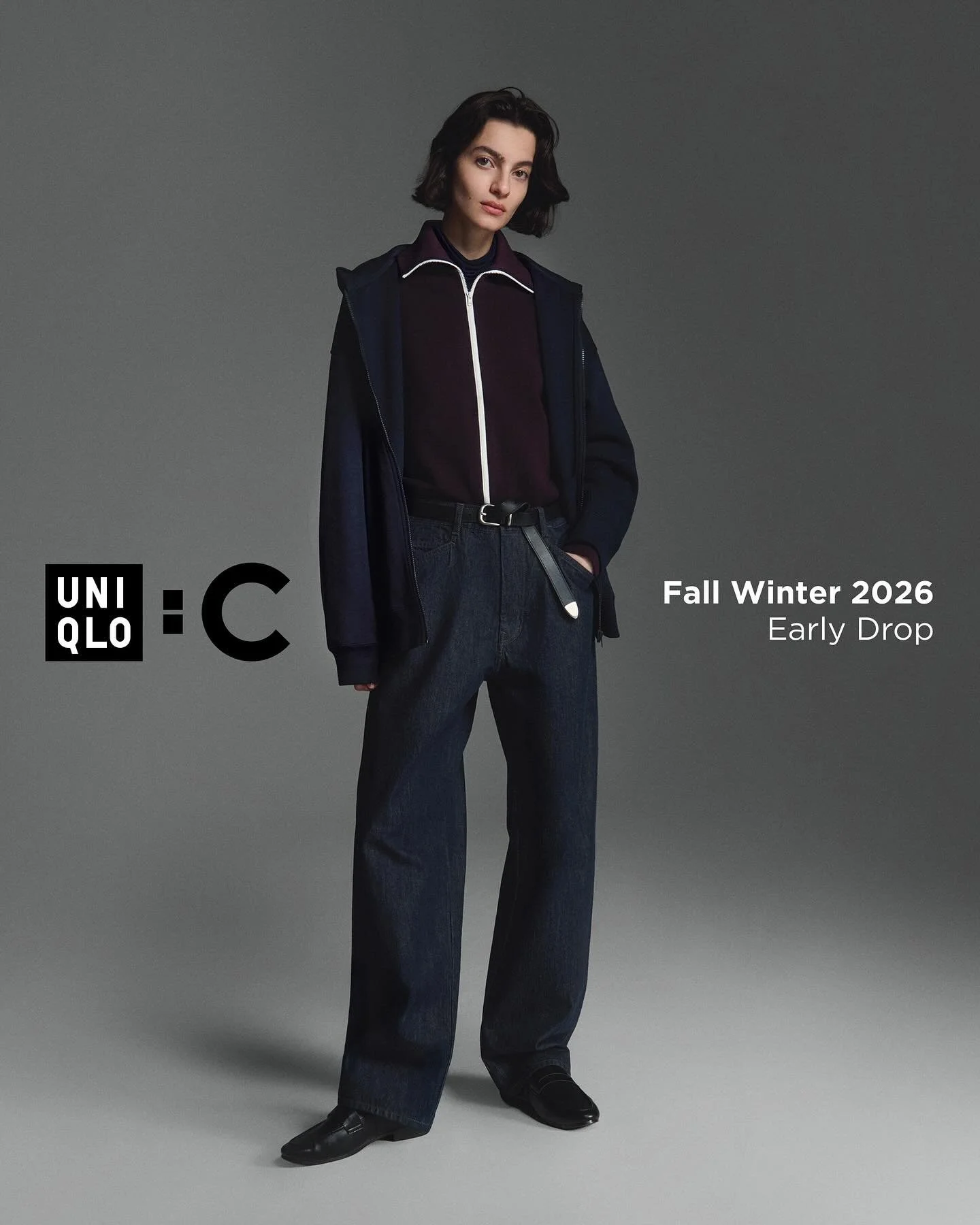 Fashion model with short dark hair wearing a dark jacket over a zip-up top and wide-leg jeans, standing against a plain gray background, promoting UNIQLO Fall Winter 2026 early drop.