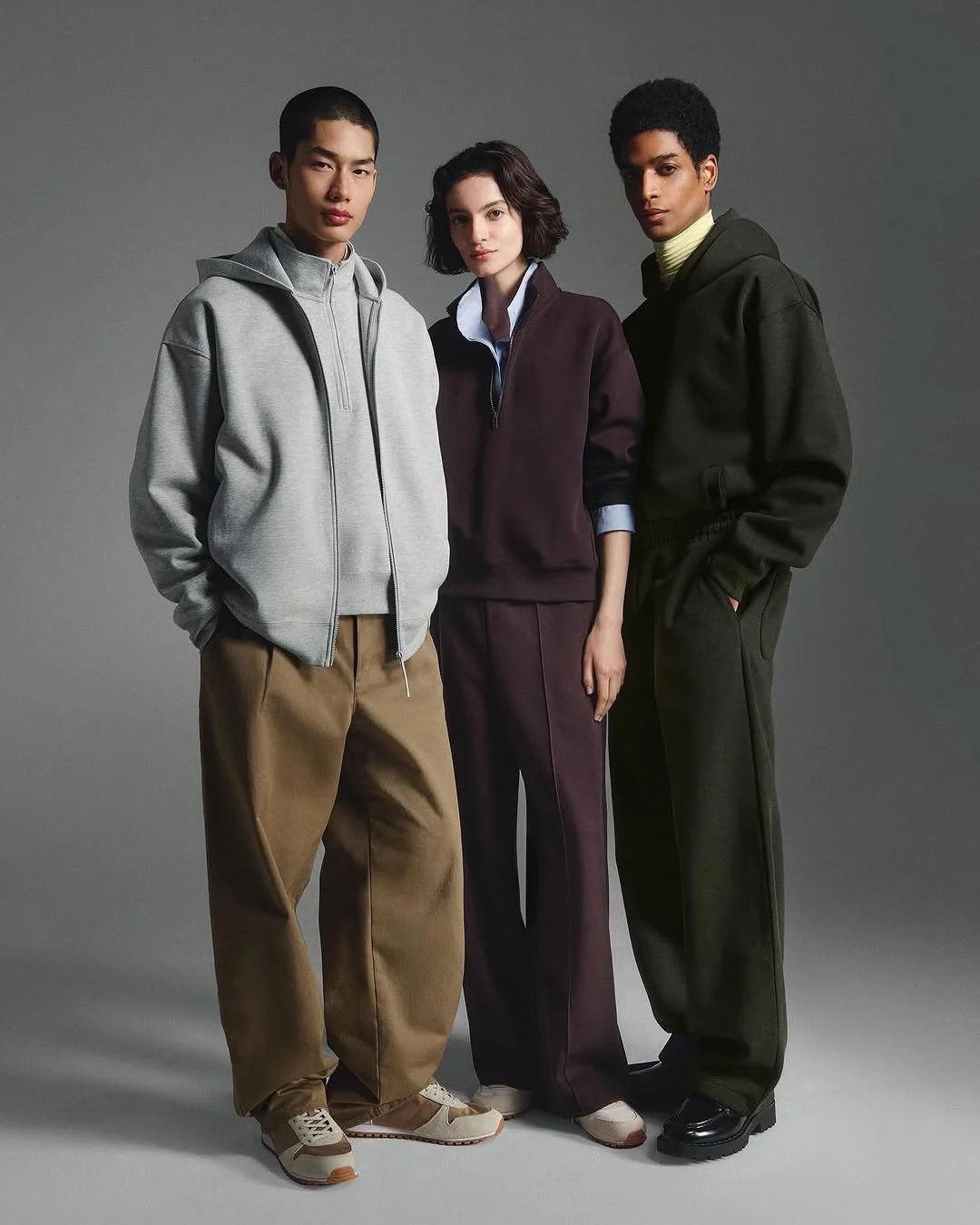 Three young adults standing together against a gray background, wearing casual streetwear with hoodies and loose pants.