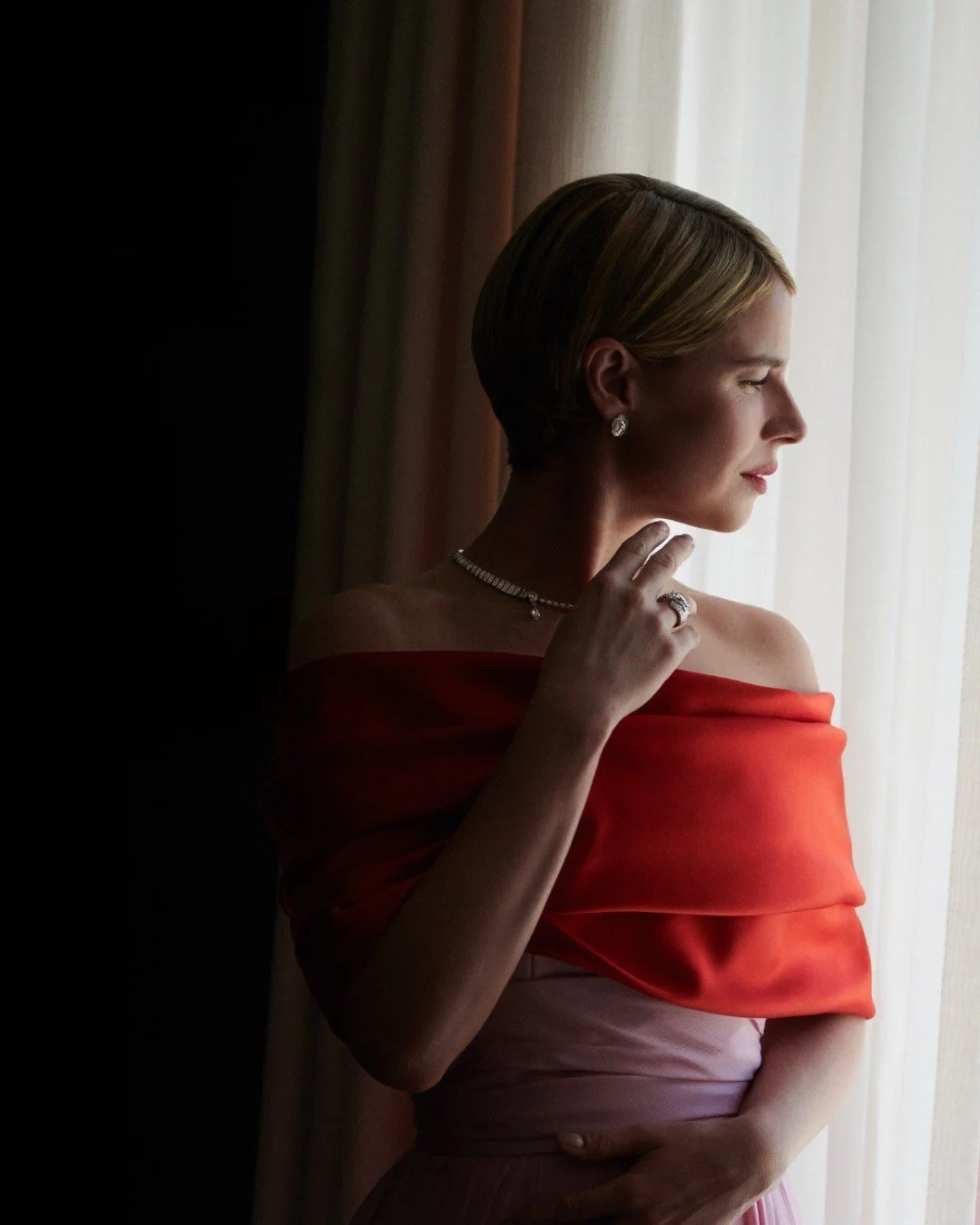 A woman with short blonde hair looking out a window, wearing a strapless red dress and silver jewelry.