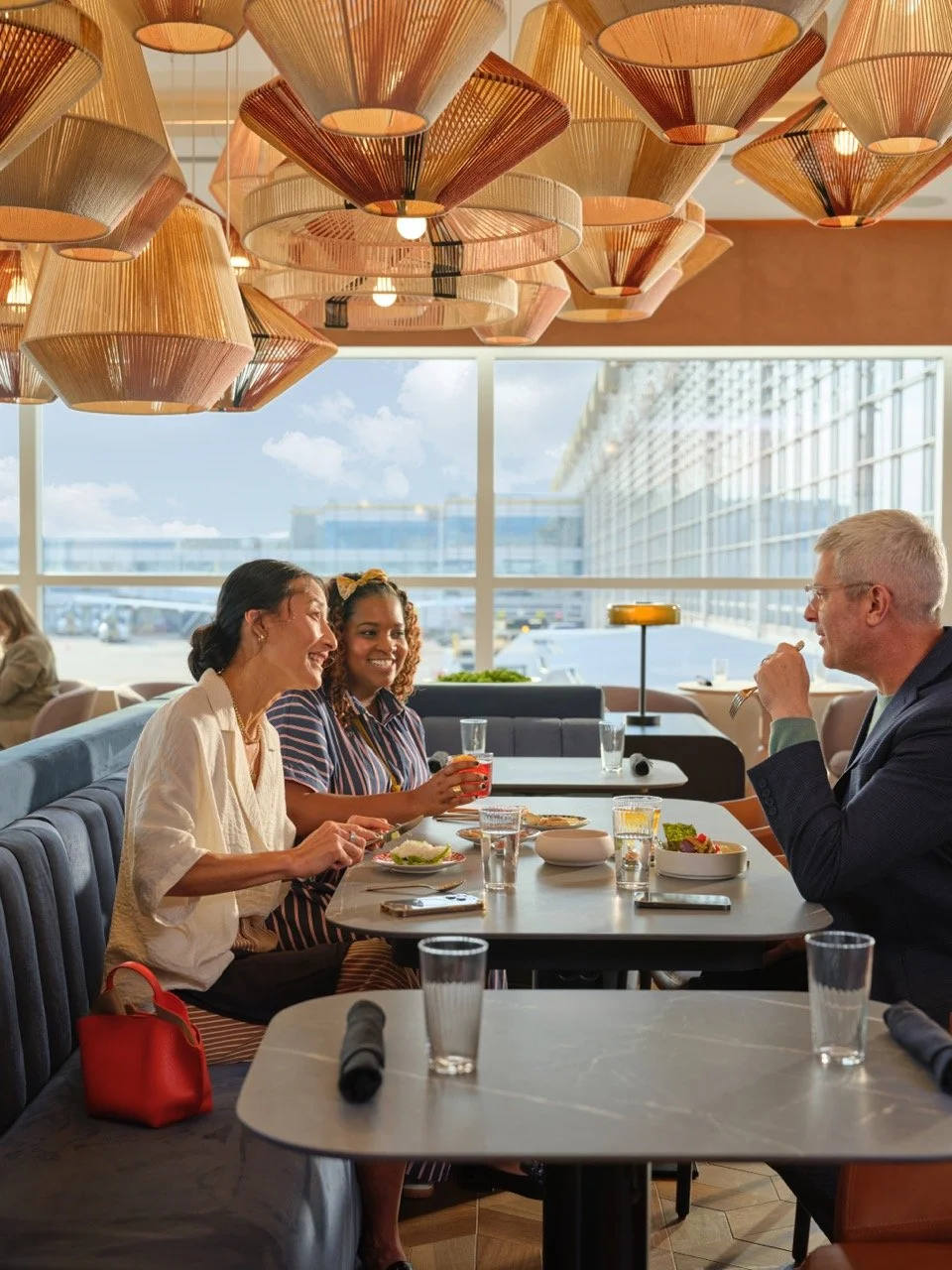 Three people sitting at a table in a restaurant, smiling and talking. Two women and one man, with food and drinks on the table, large windows behind them showing a building and blue sky, with decorative hanging lights above.