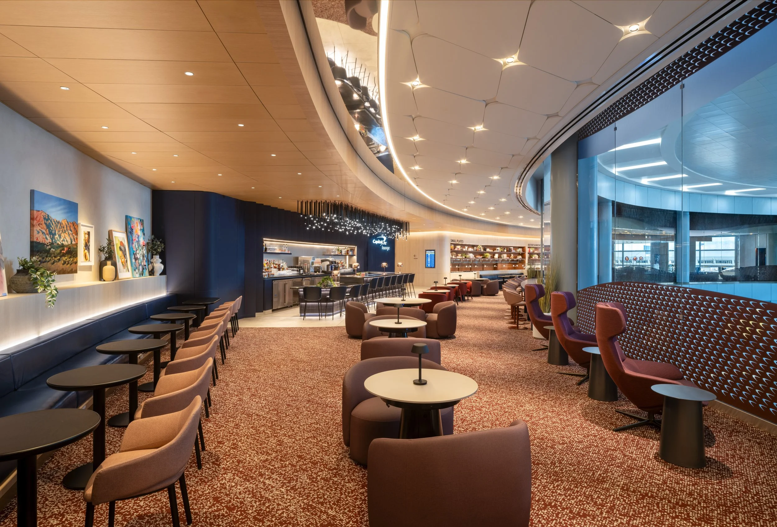 Modern lounge area with a curved ceiling, artwork on the wall, bar area, and seating arrangements with chairs and tables.