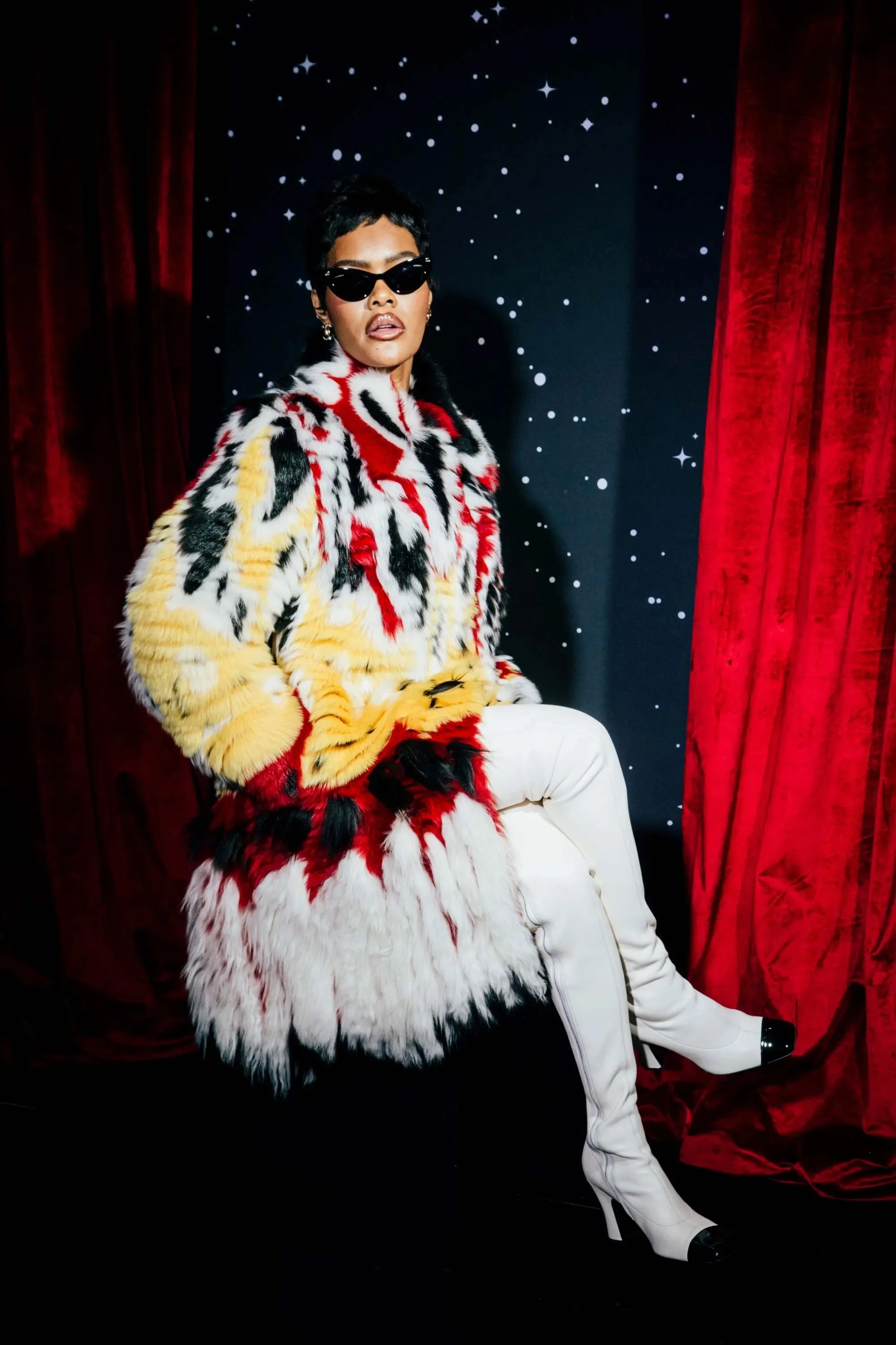 A woman with short dark hair wearing sunglasses and a colorful fur coat, sitting in front of a dark backdrop with white star-like patterns, red velvet curtains on the sides, and white pants with high heels.