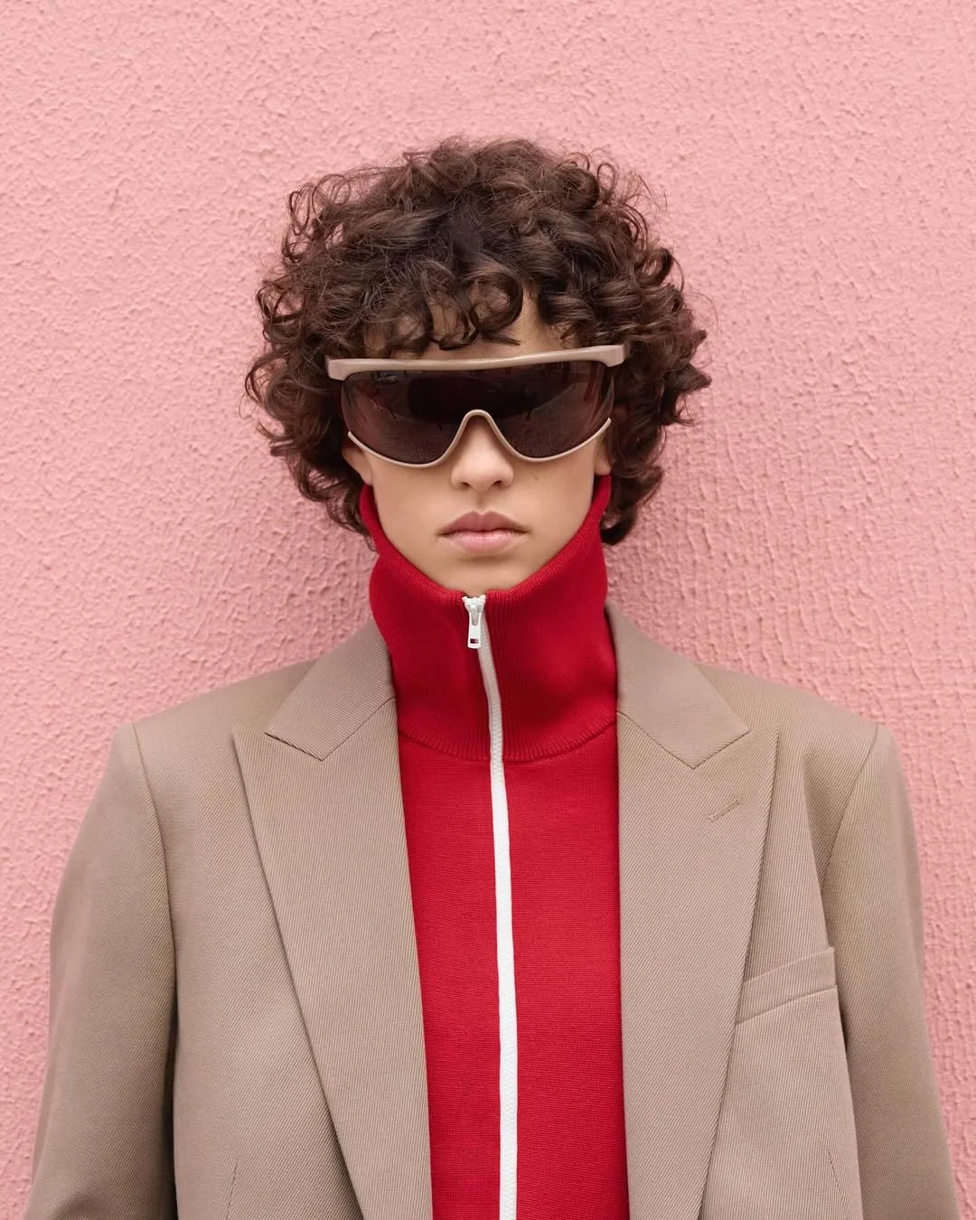 Person with curly hair wearing large sunglasses, a beige blazer, and a red zip-up jacket, standing against a pink textured wall.