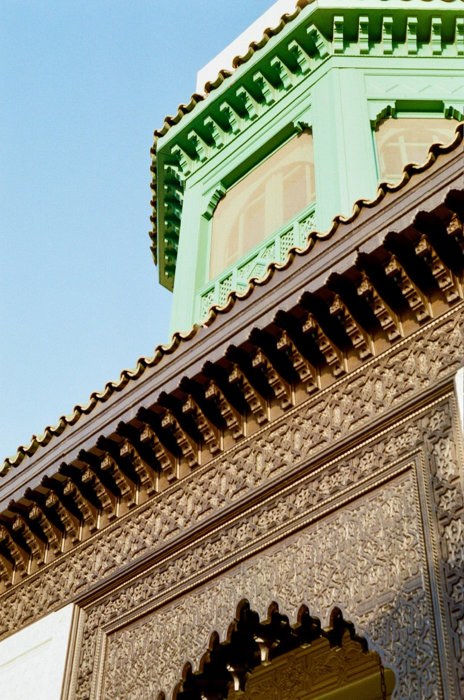 Close-up of a decorated architectural corner with ornate detailing, wooden latticework, and a green structure on the upper part against a clear blue sky backdrop.