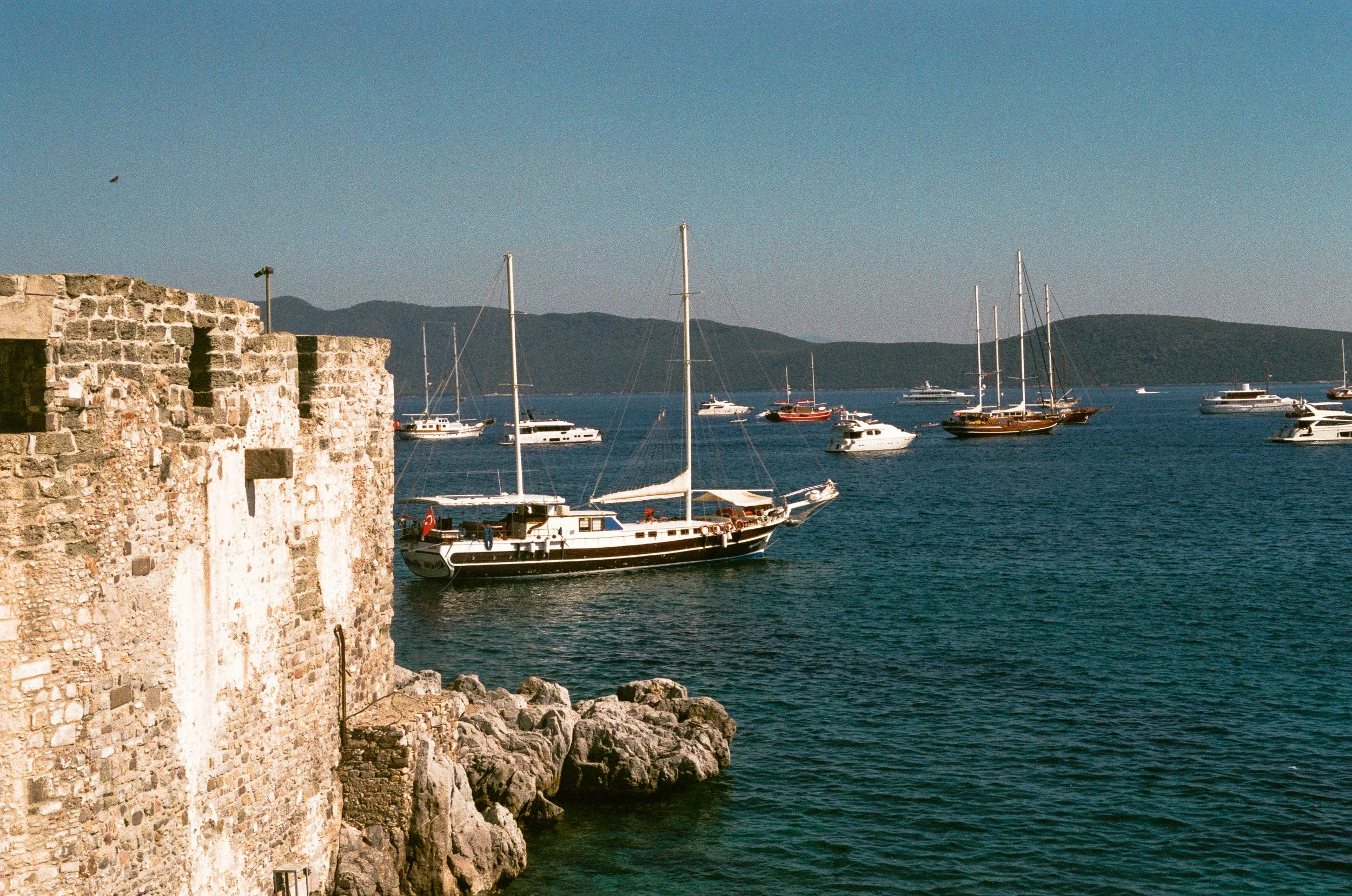Boats anchored in a harbor next to a stone wall with hills in the background under a clear blue sky.