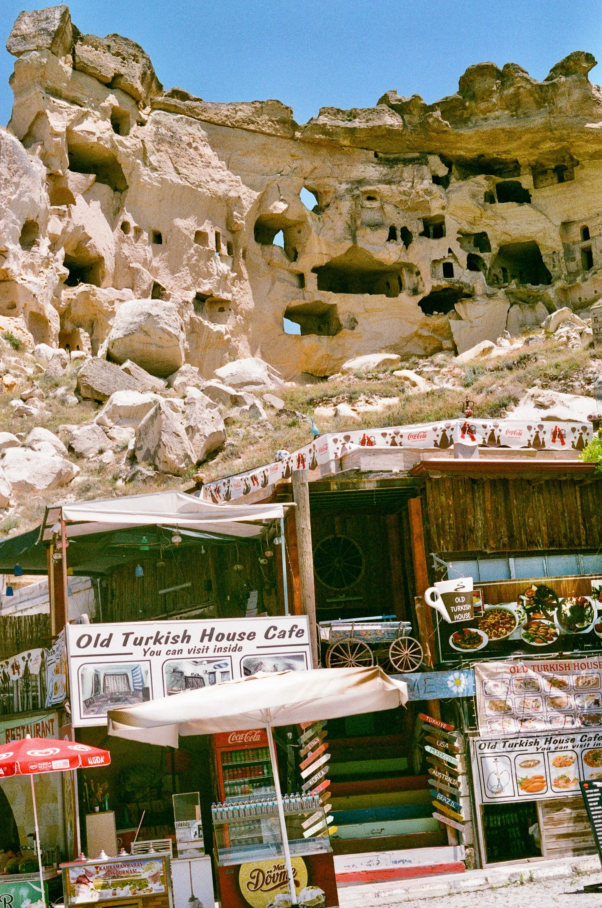 Outdoor stall selling Turkish food and souvenirs, with a rocky cliff face in the background under a clear blue sky.