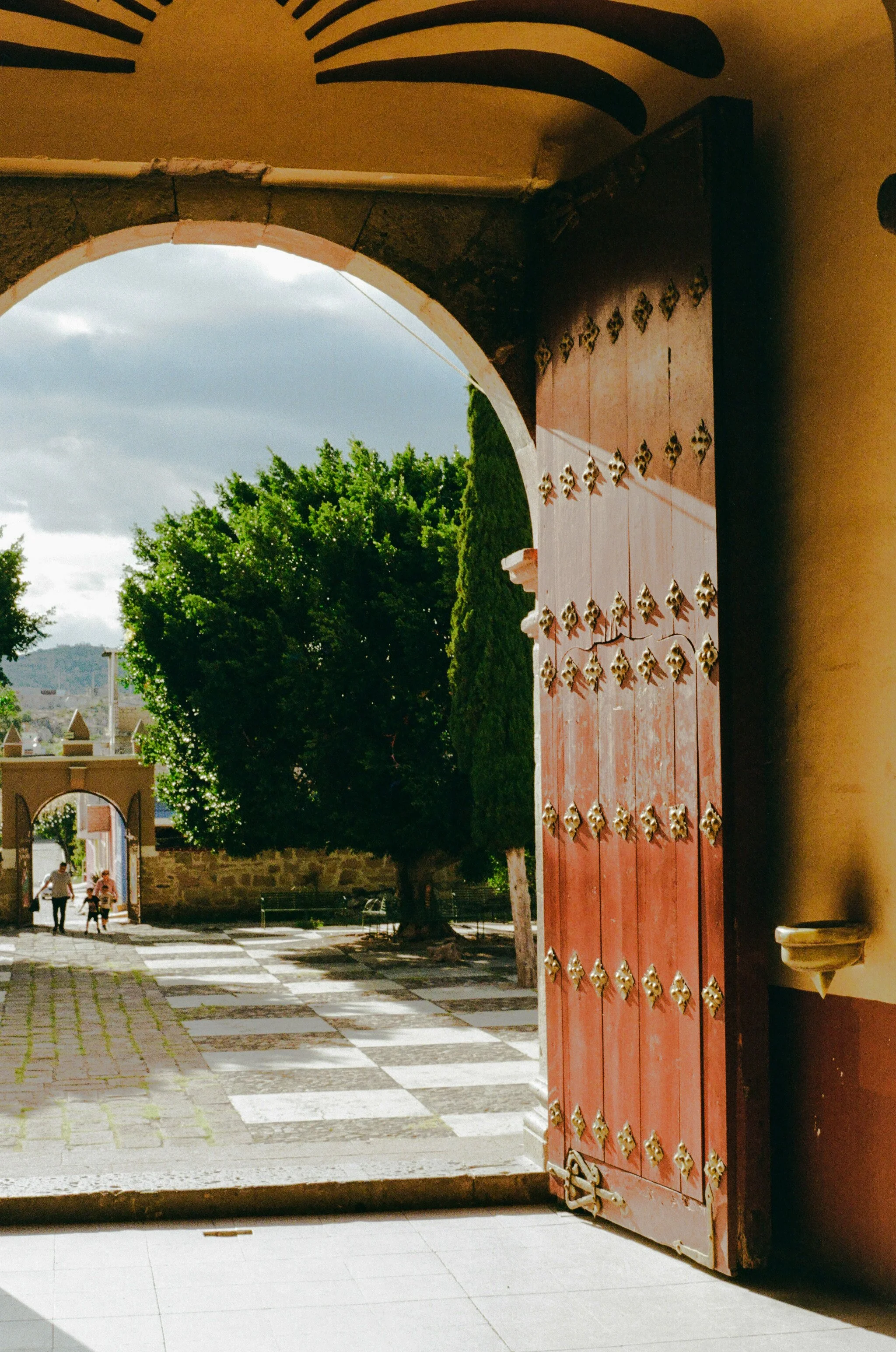An arched doorway leading to a courtyard with a checkered tile pathway, trees, and a stone wall, with a large wooden door on the right side decorated with metal accents.
