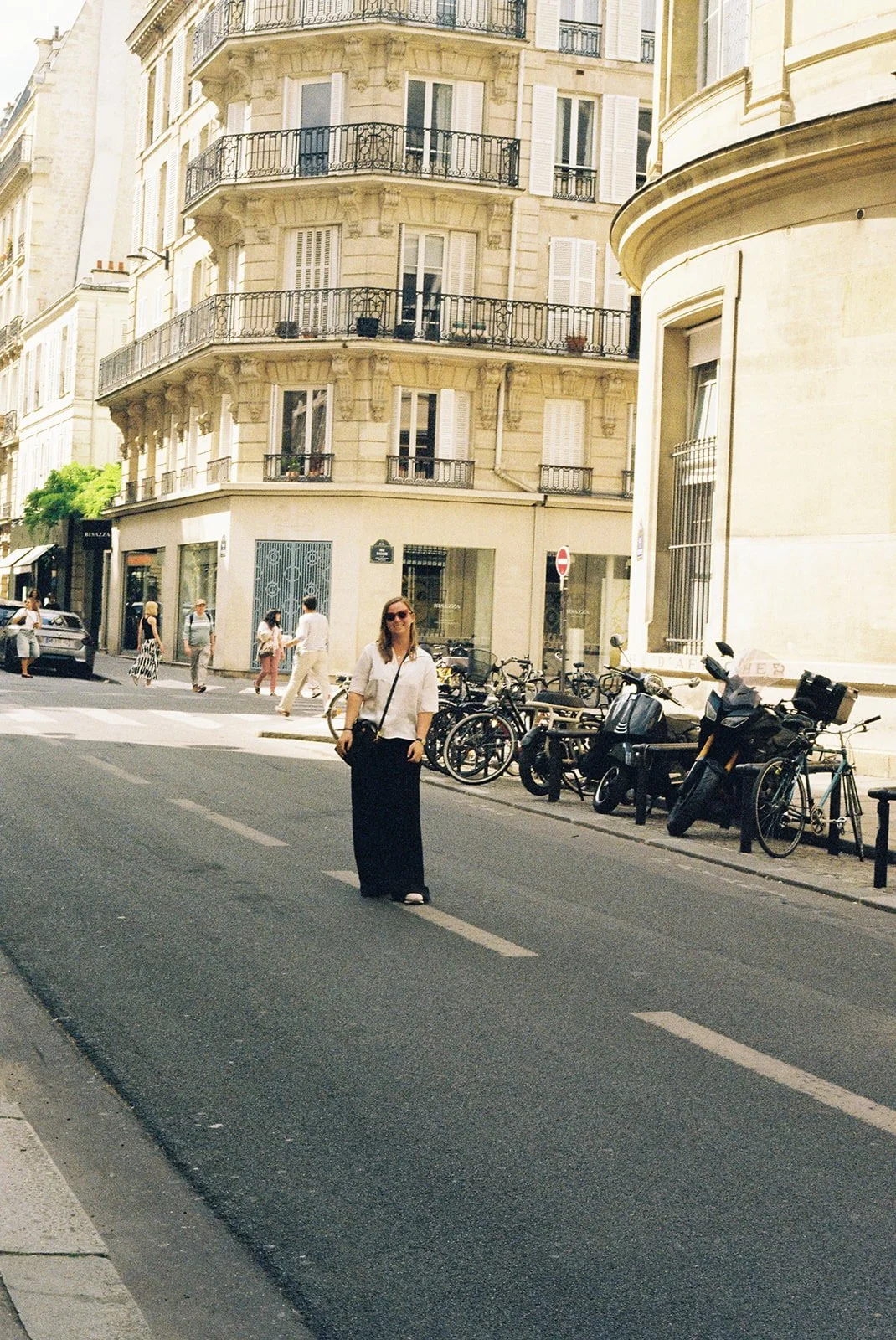 A woman standing in the middle of a city street in front of Parisian-style buildings with balconies. Several bicycles are parked along the sidewalk, and other pedestrians walk across the street.