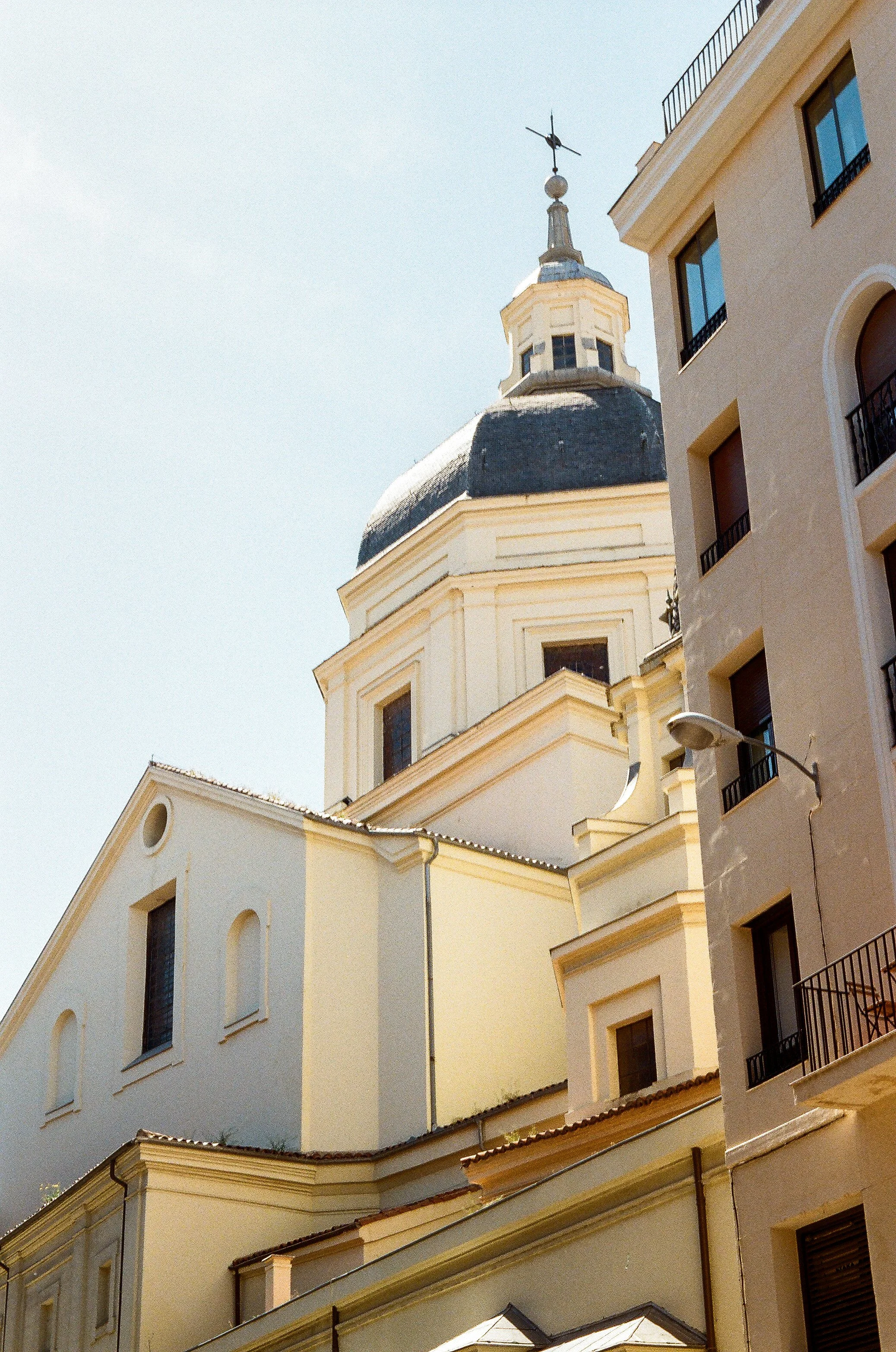 Close-up view of historic European-style buildings with beige and white facades, multiple windows with dark shutters and black wrought-iron balconies, and a church steeple with a clock and weather vane against a clear blue sky.