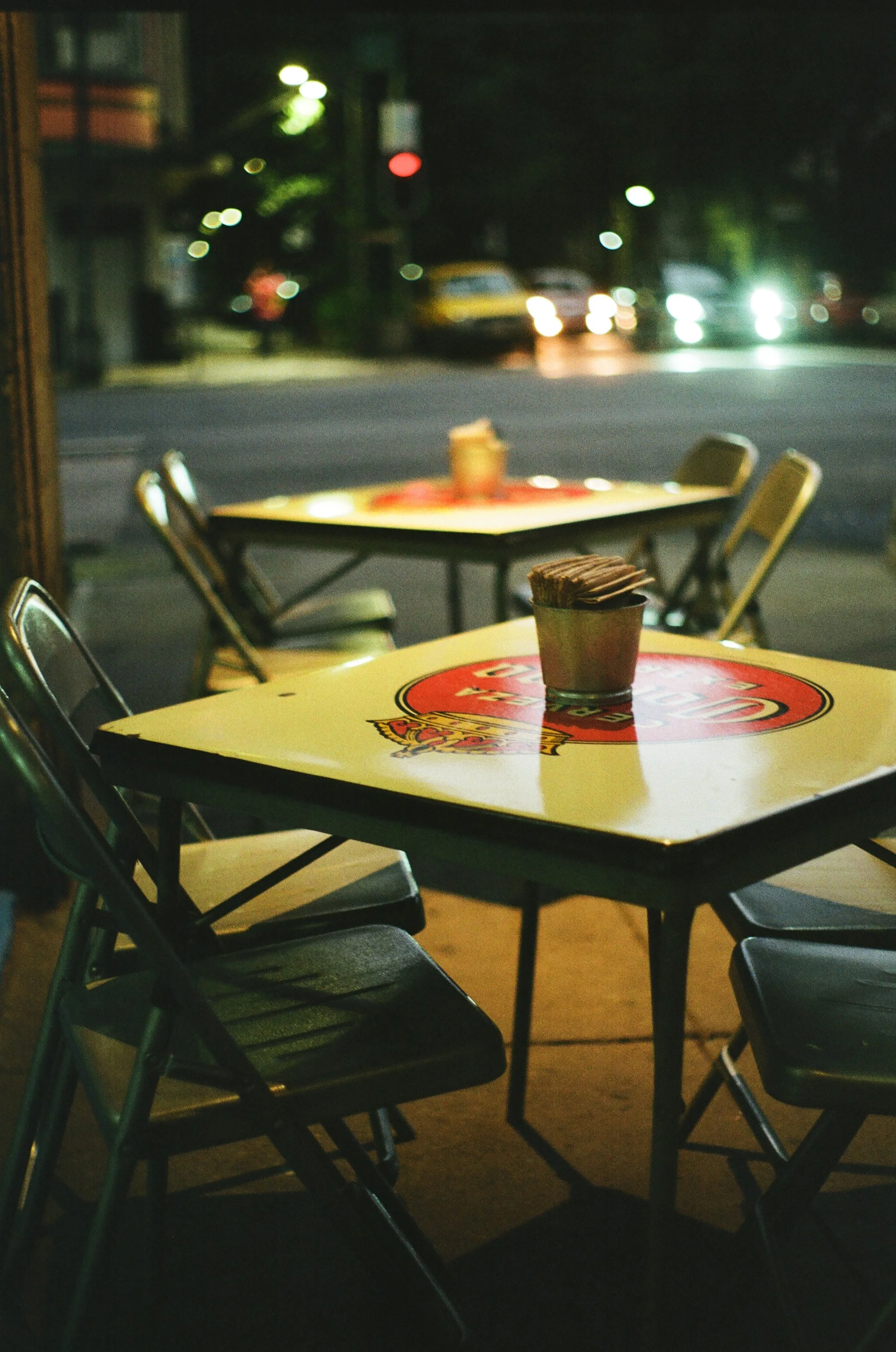 Nighttime scene of two empty yellow tables with chairs outside a restaurant, one with a small potted plant, and street with cars and blurred lights in the background.