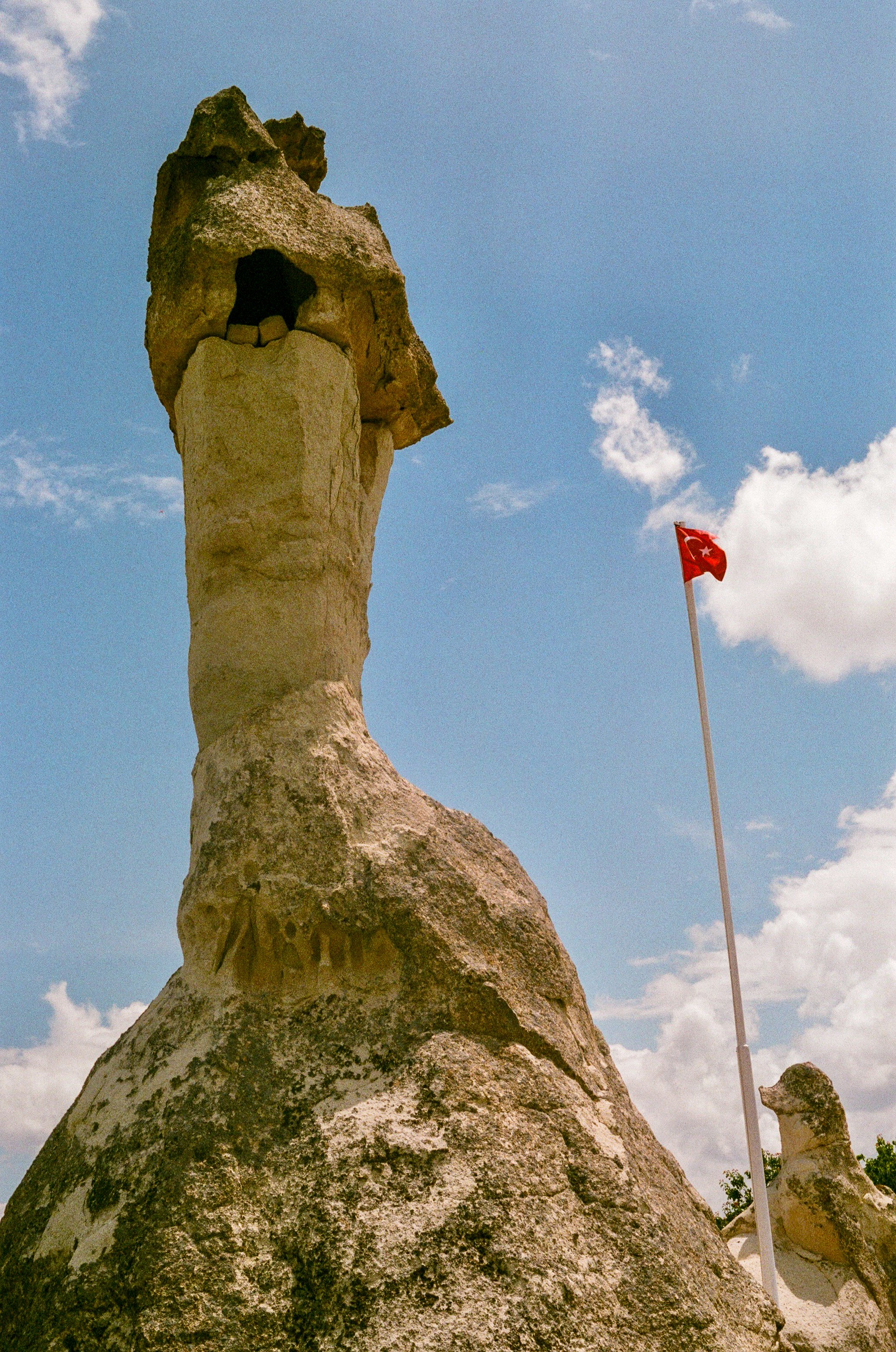 Large rock formation with carved inscription, Turkish flag on pole, partly cloudy sky.