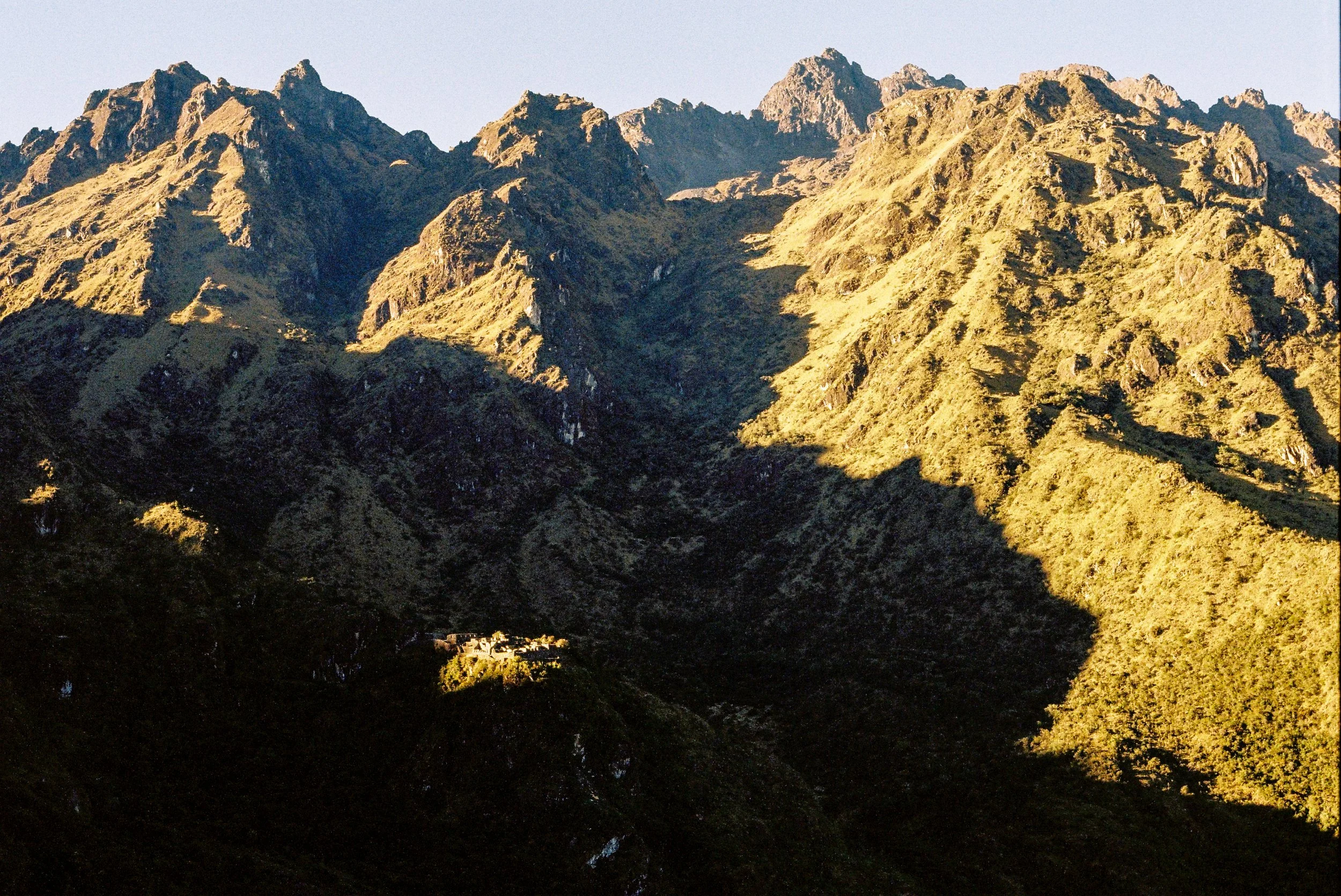 Mountains with rugged peaks and slopes, illuminated by sunlight, with a small house or building at the base surrounded by trees.