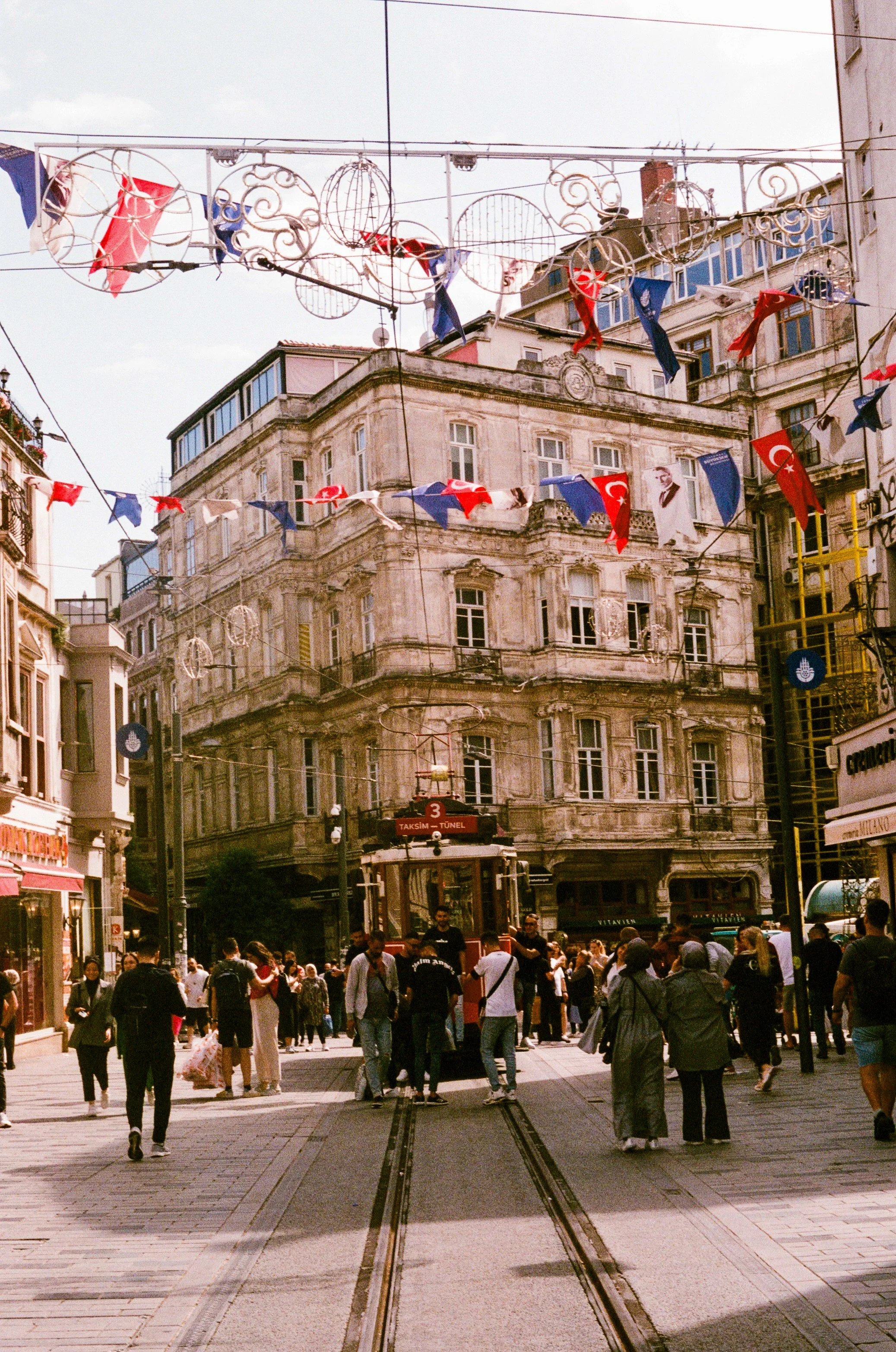 A busy street scene in Istanbul, Turkey, with pedestrians walking on the street, trams, and historic buildings, decorated with flags and circular light fixtures hanging above.