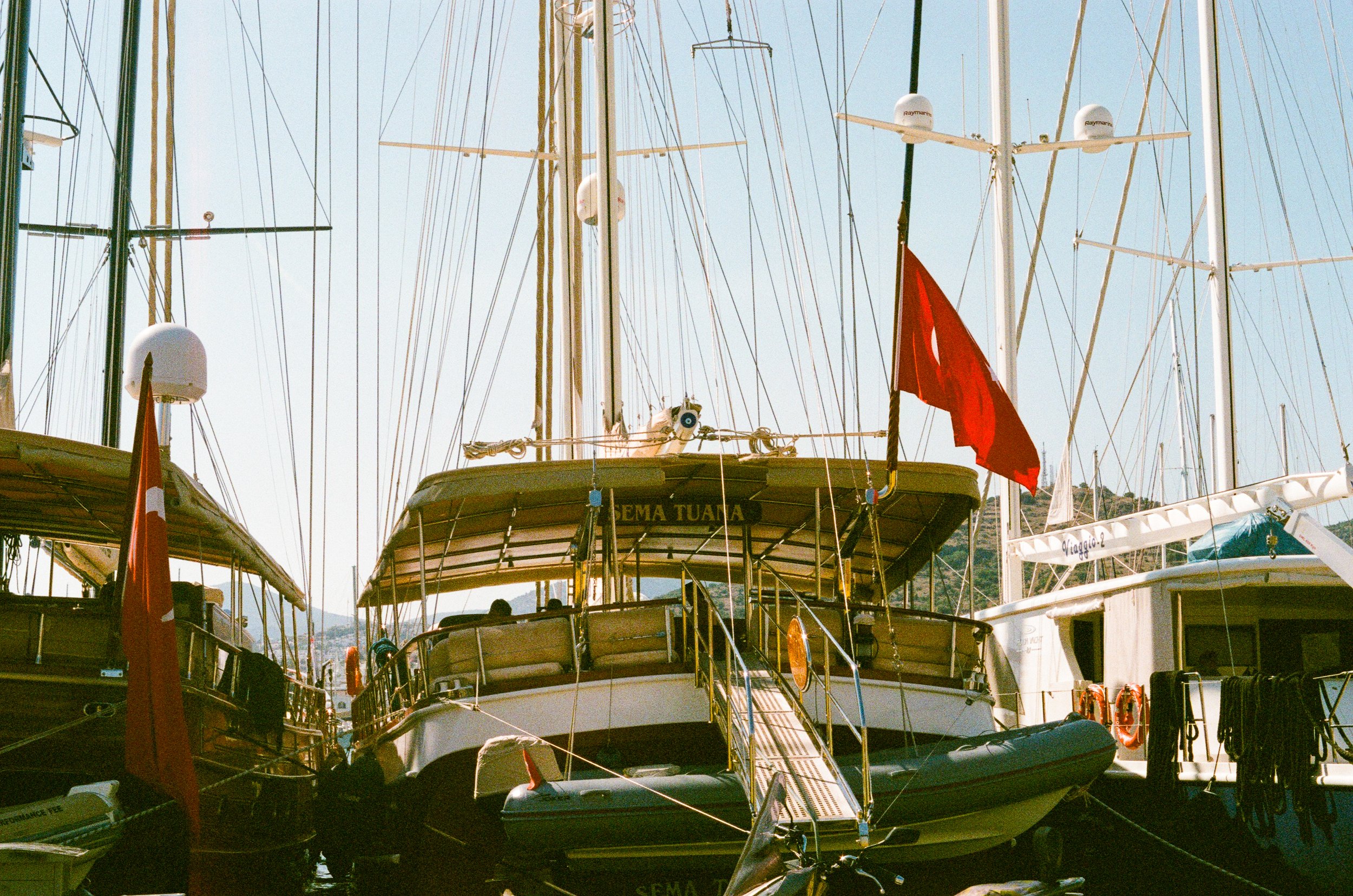 A marina filled with sailboats docked side by side, with masts and rigging visible against a clear sky. Some boats have visible name tags and flags.