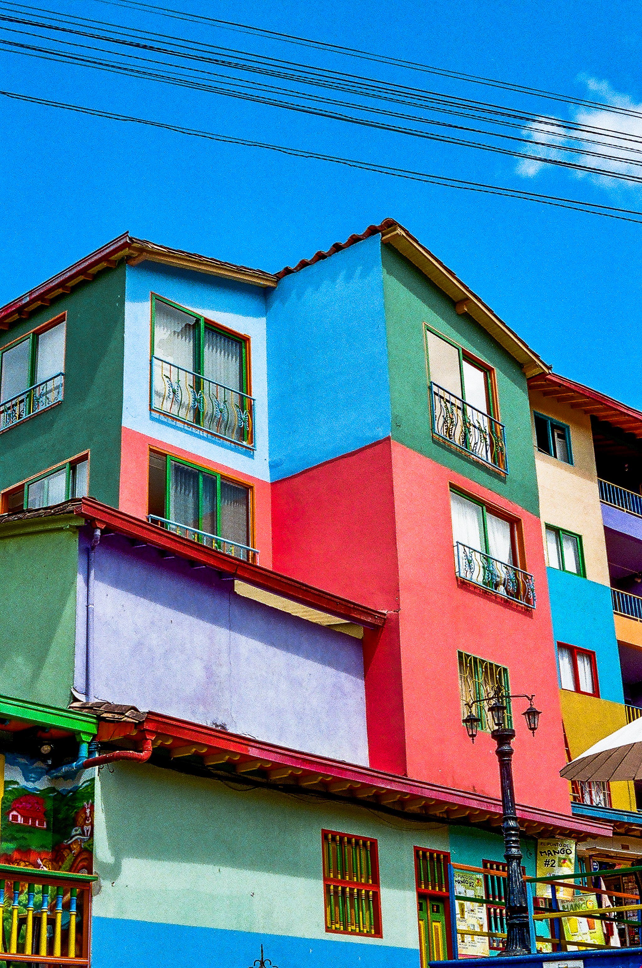 Colorful building with multiple stories painted in various vibrant colors including green, blue, red, and yellow, with balconies and windows. There is a clear blue sky with power lines overhead and a street lamp in the foreground.