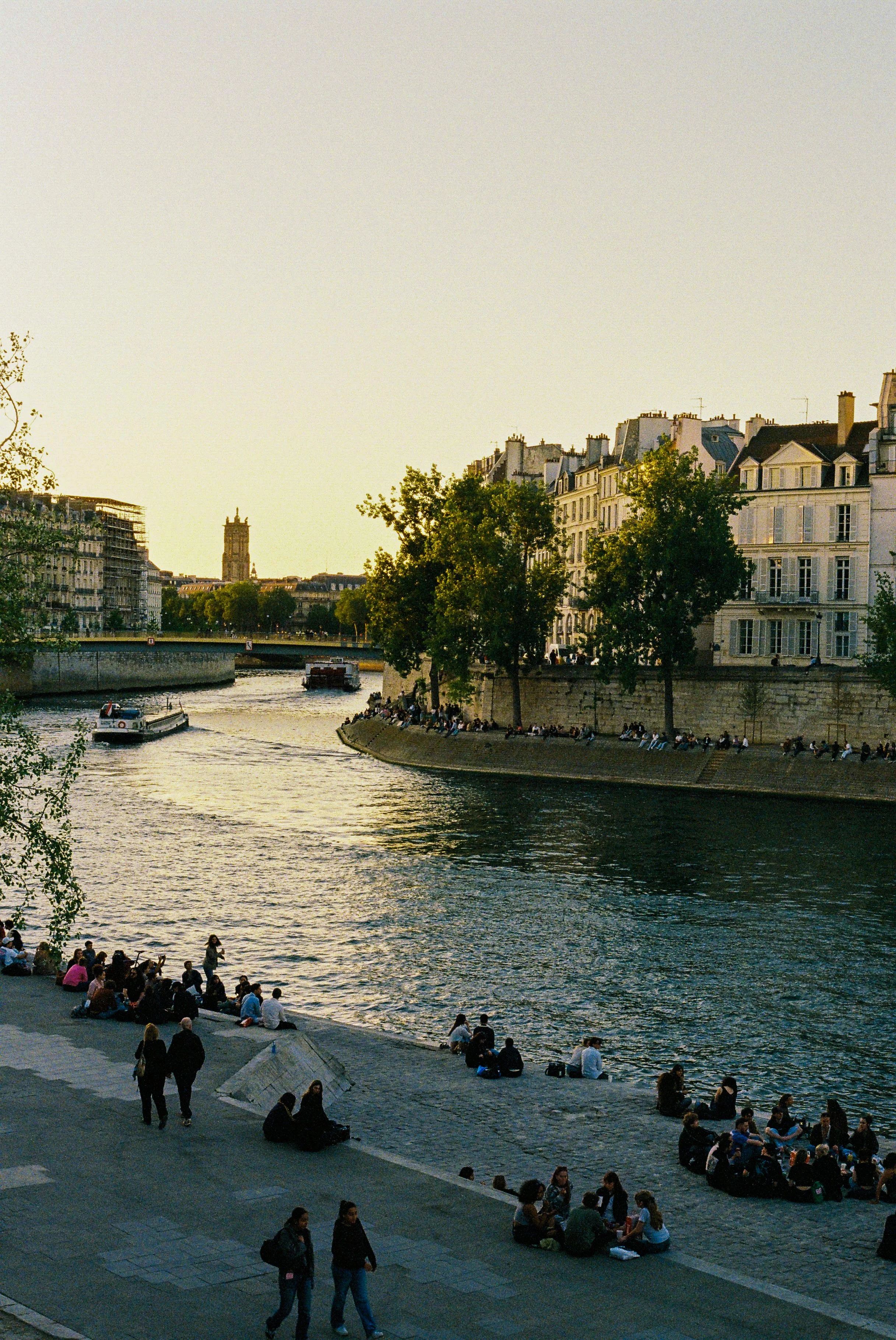 People sitting and walking along the riverbank at sunset in a city with historic buildings and a church tower in the background.