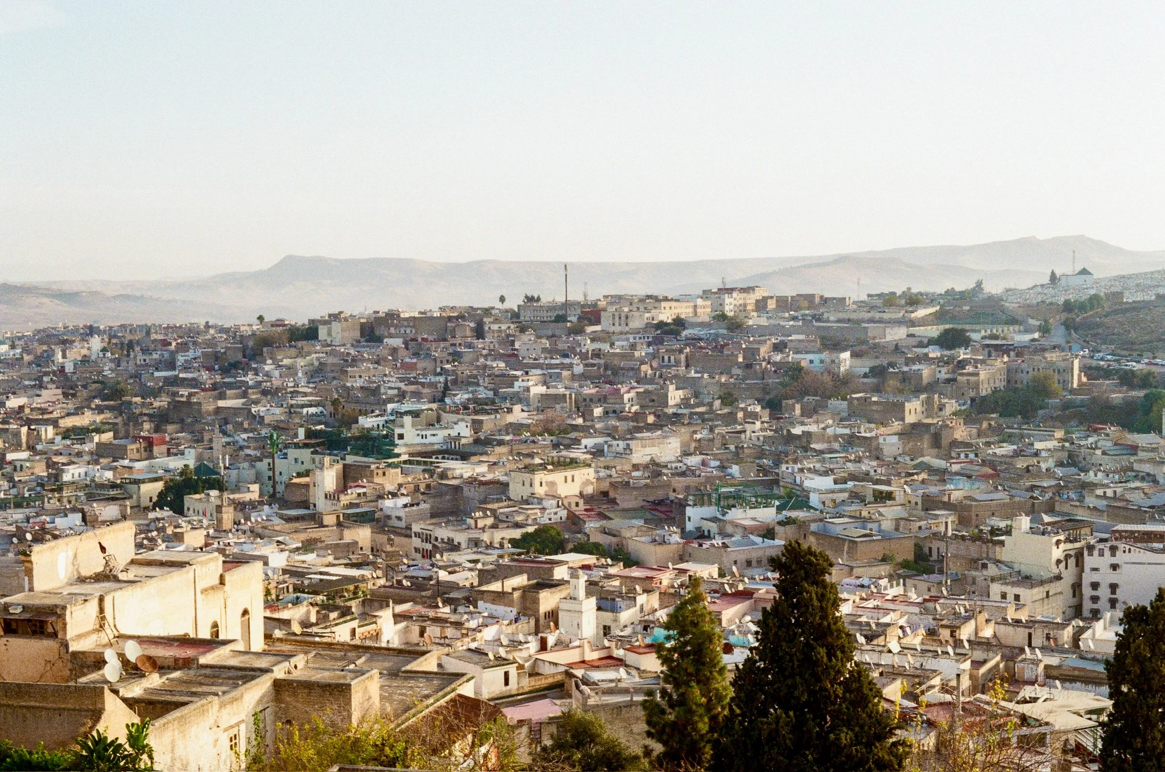 A cityscape with numerous densely packed buildings, some with flat rooftops, and a hill with sparse vegetation in the background under a clear sky.