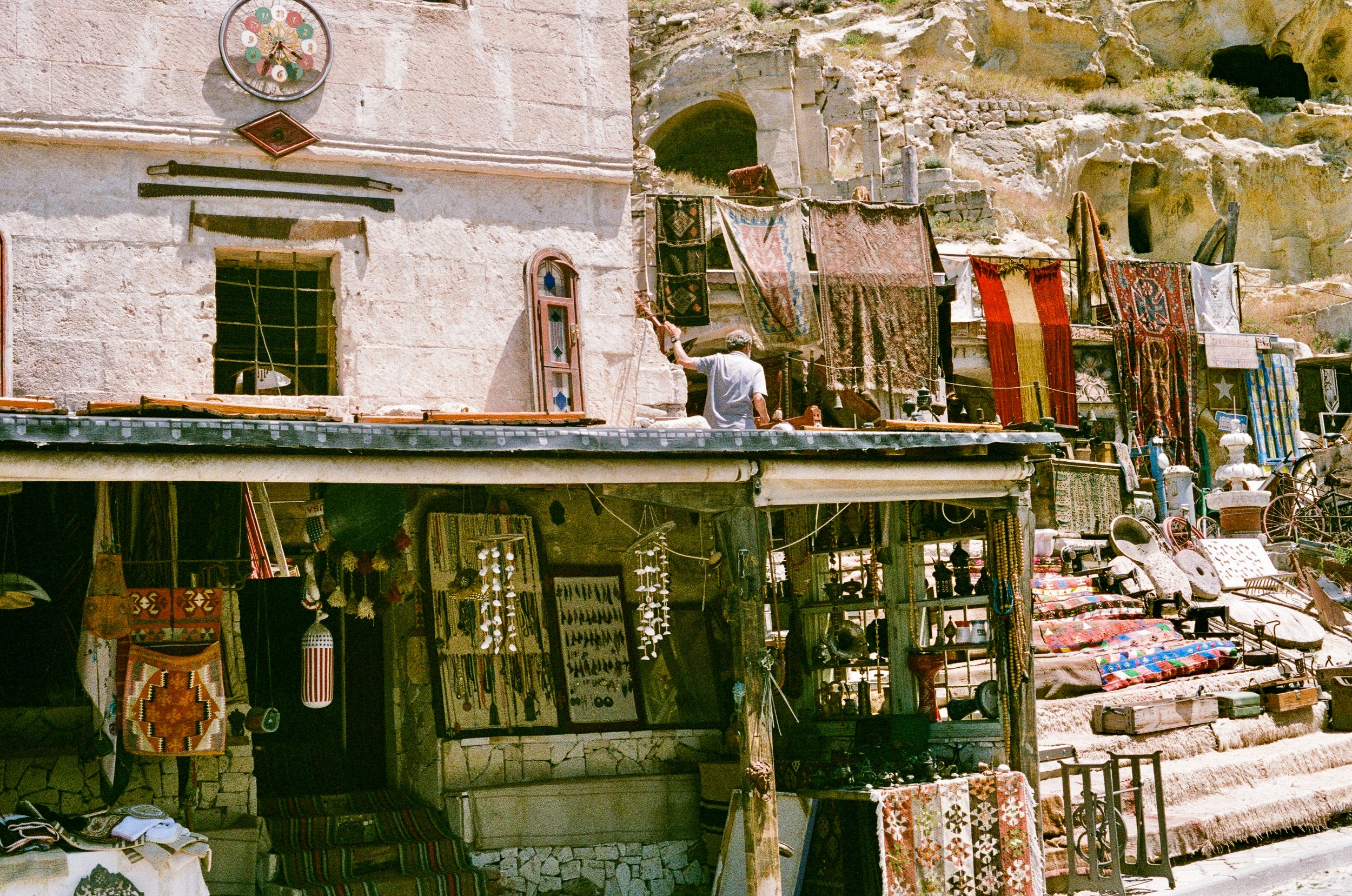 A street market with handmade textiles and crafts displayed on shops and hanging racks, behind a stone building with a clock, set against a rocky hillside with caves.