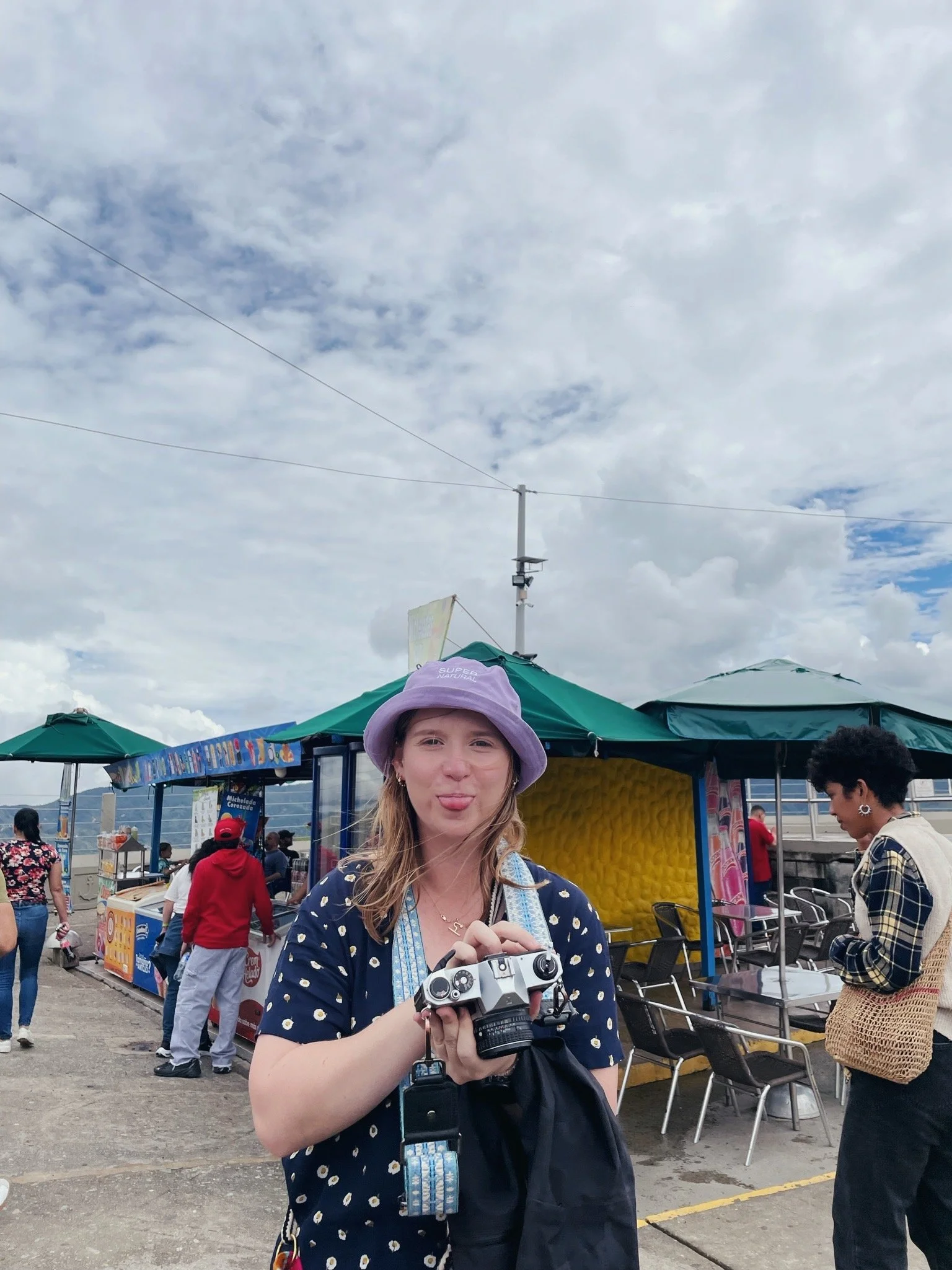 A woman with sunglasses holding a vintage camera, standing in an outdoor market with umbrellas and people in the background.