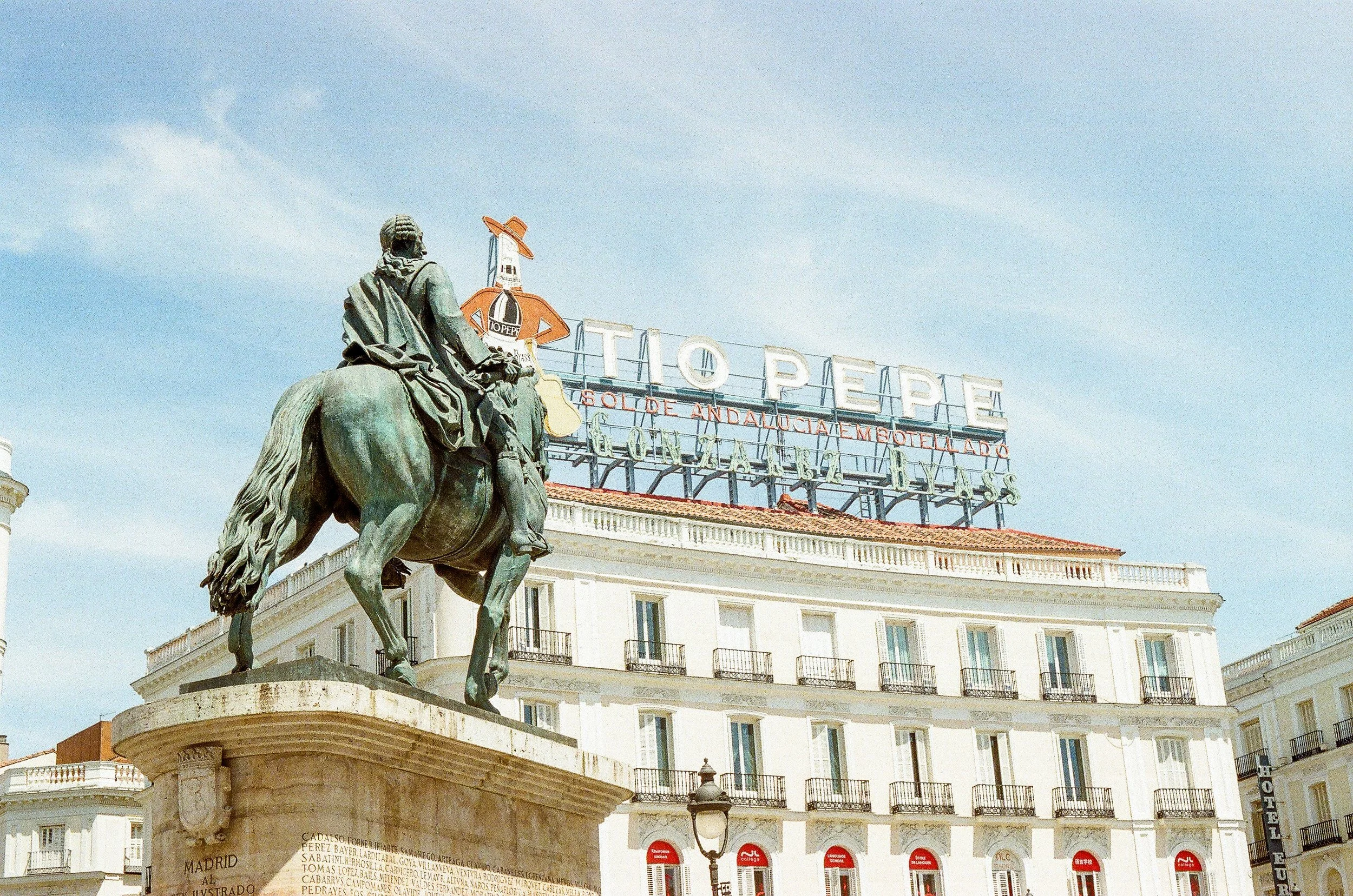 A statue of a man on a horse in front of a large building with a sign that reads 'TOPEPE.' The statue is on a pedestal with inscriptions, and rooftops and windows of the building are visible below the sign.