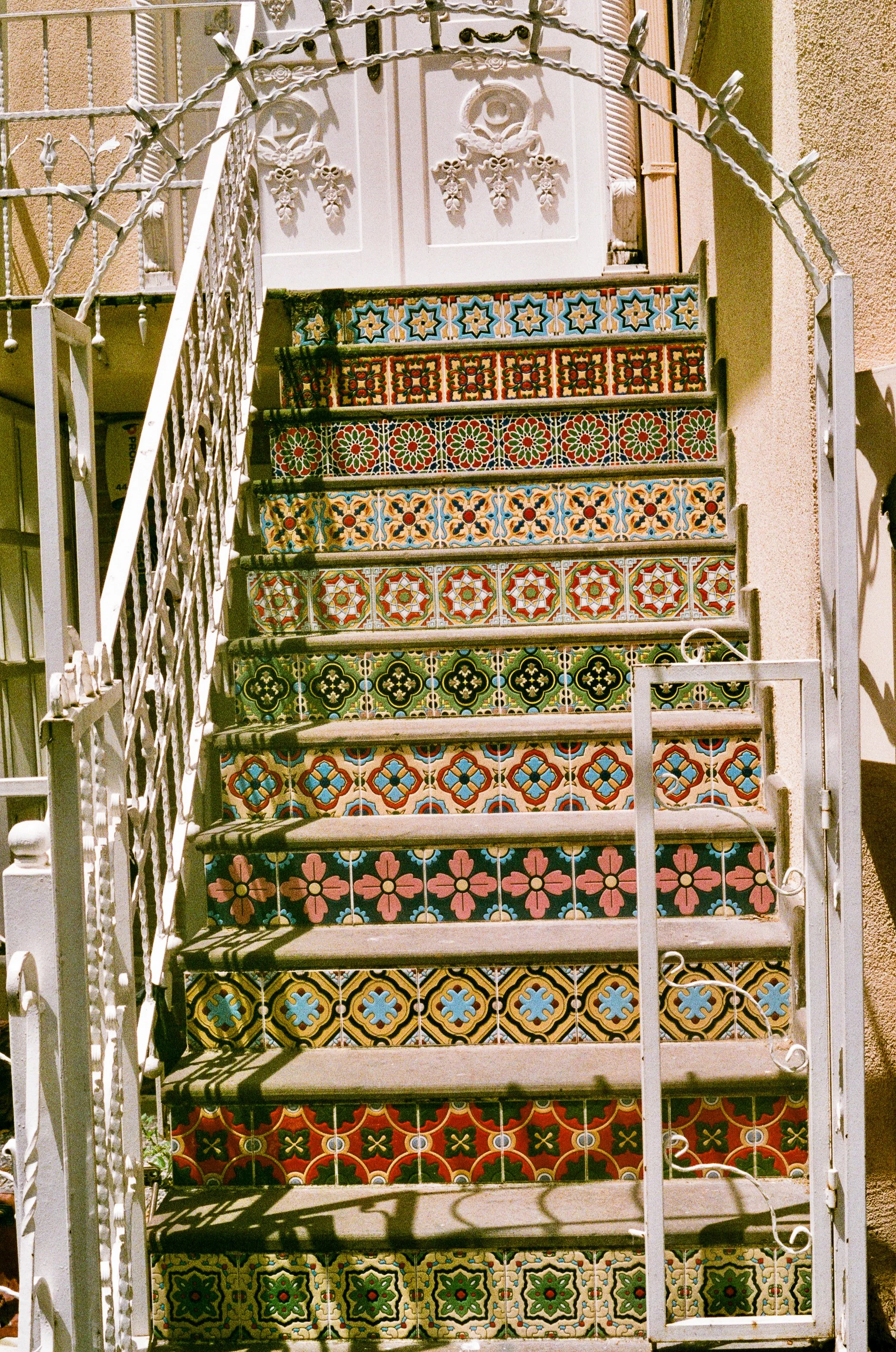 A staircase with colorful decorative tiles on each step leading to a white door with ornate details. The staircase has white wrought iron railings and shadows cast on the steps from the railings.
