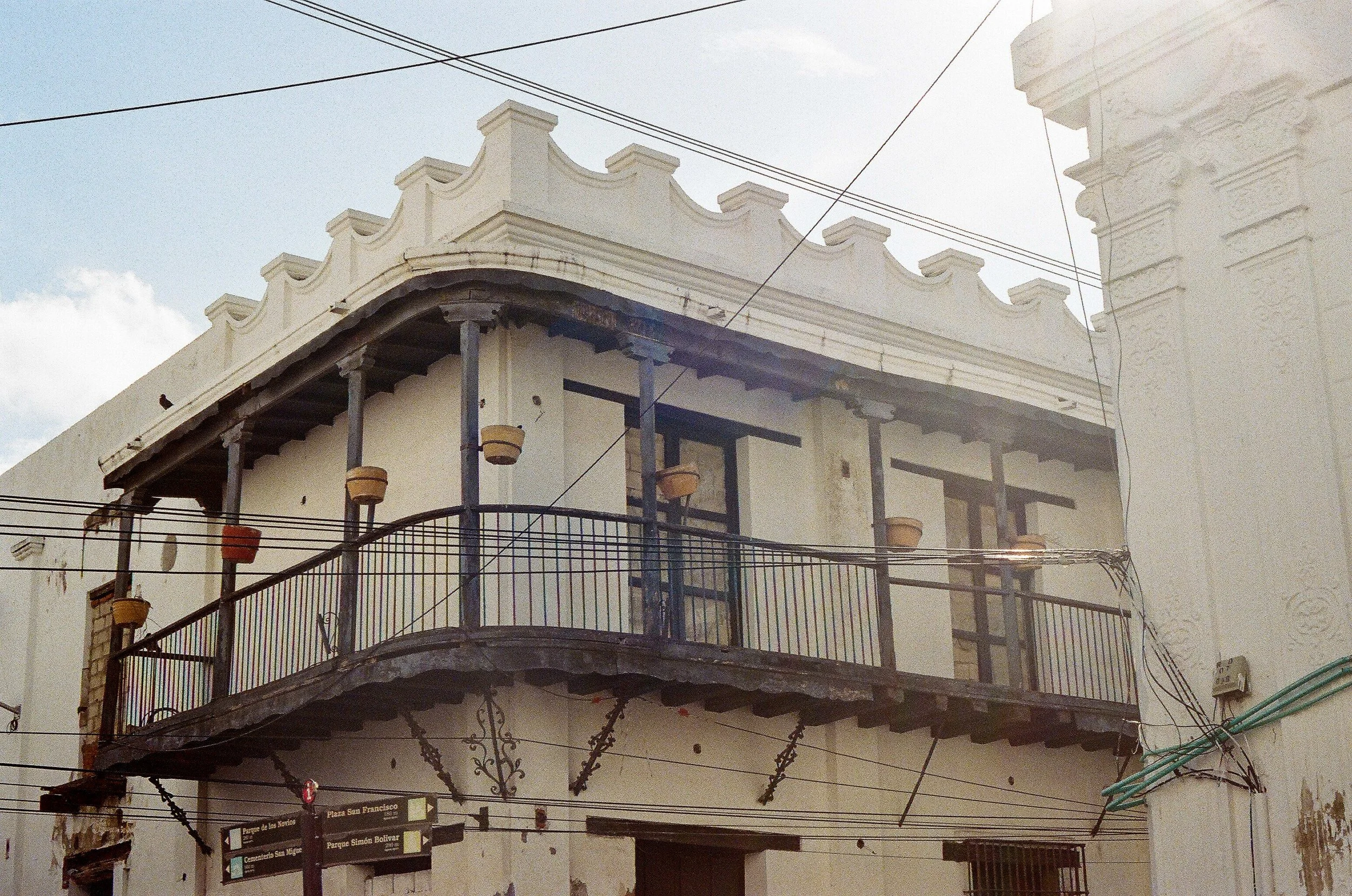 A building with traditional architectural style, featuring multiple balconies with iron railings, decorative molding along the roofline, and hanging flower pots. There are electrical wires across the image and signs at the bottom indicating nearby lo