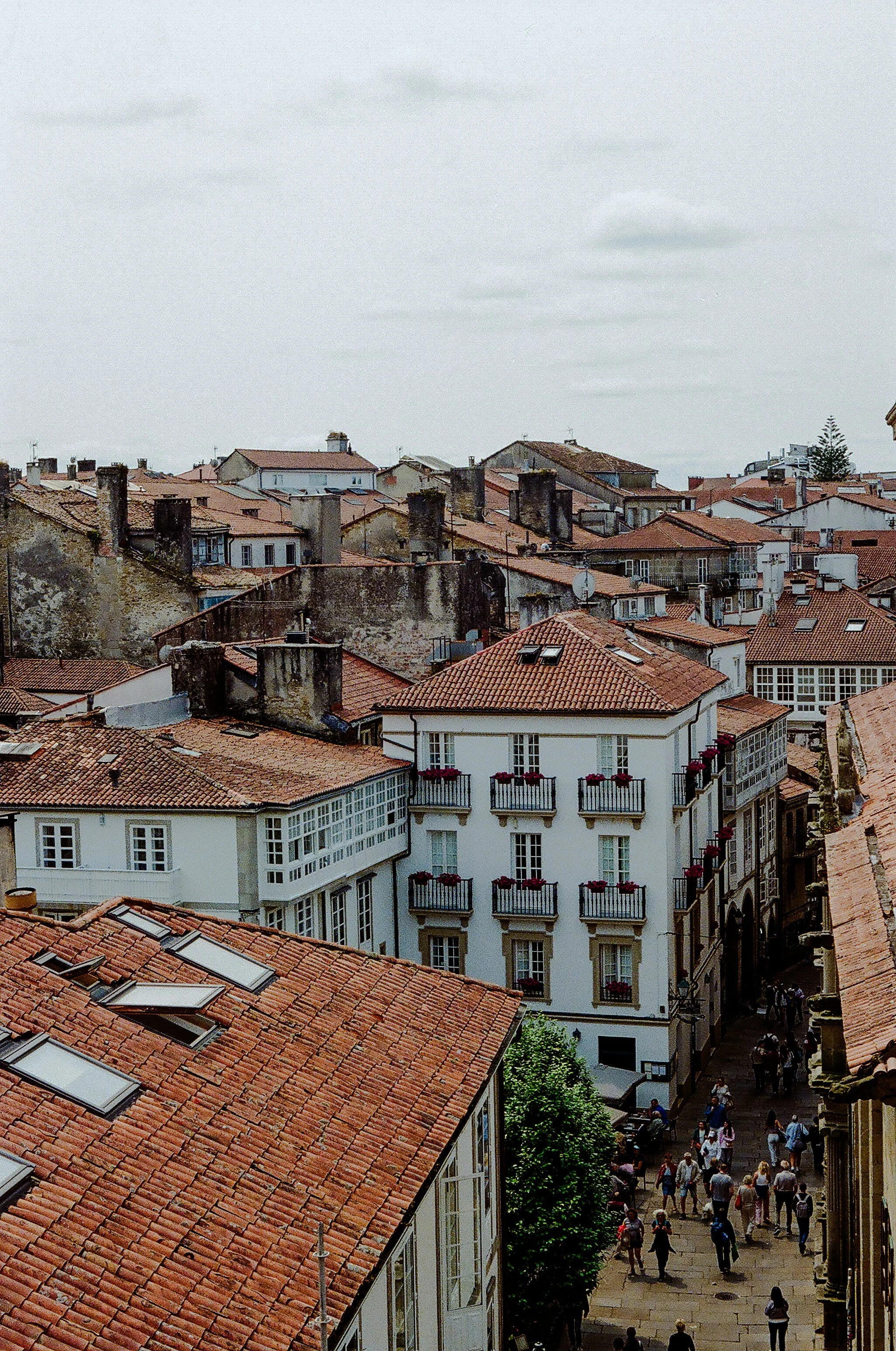A view of a city with red-tiled roofs and a narrow street filled with pedestrians.