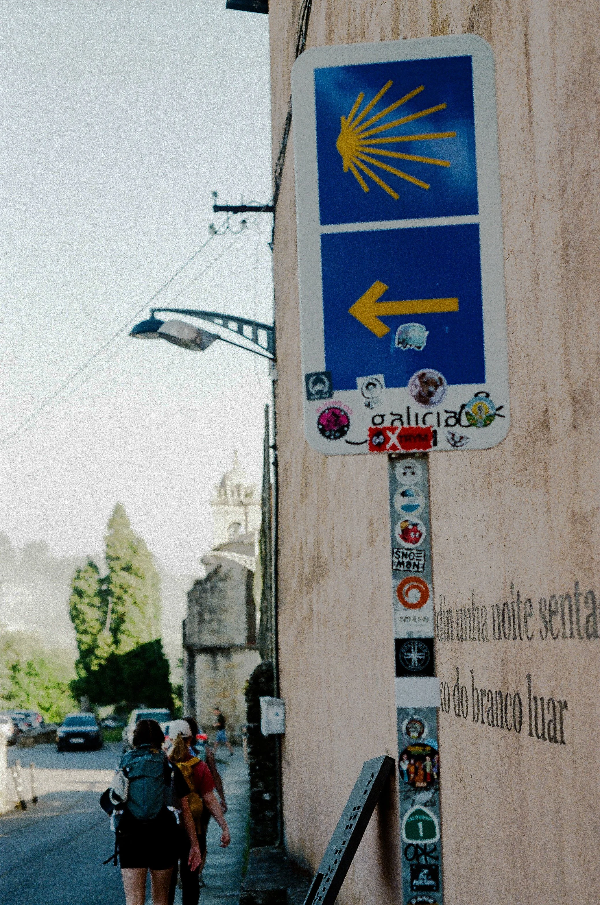 Street scene with people walking along a sidewalk, street lamps, and a wall with various stickers and graffiti; some buildings and trees visible in the background.