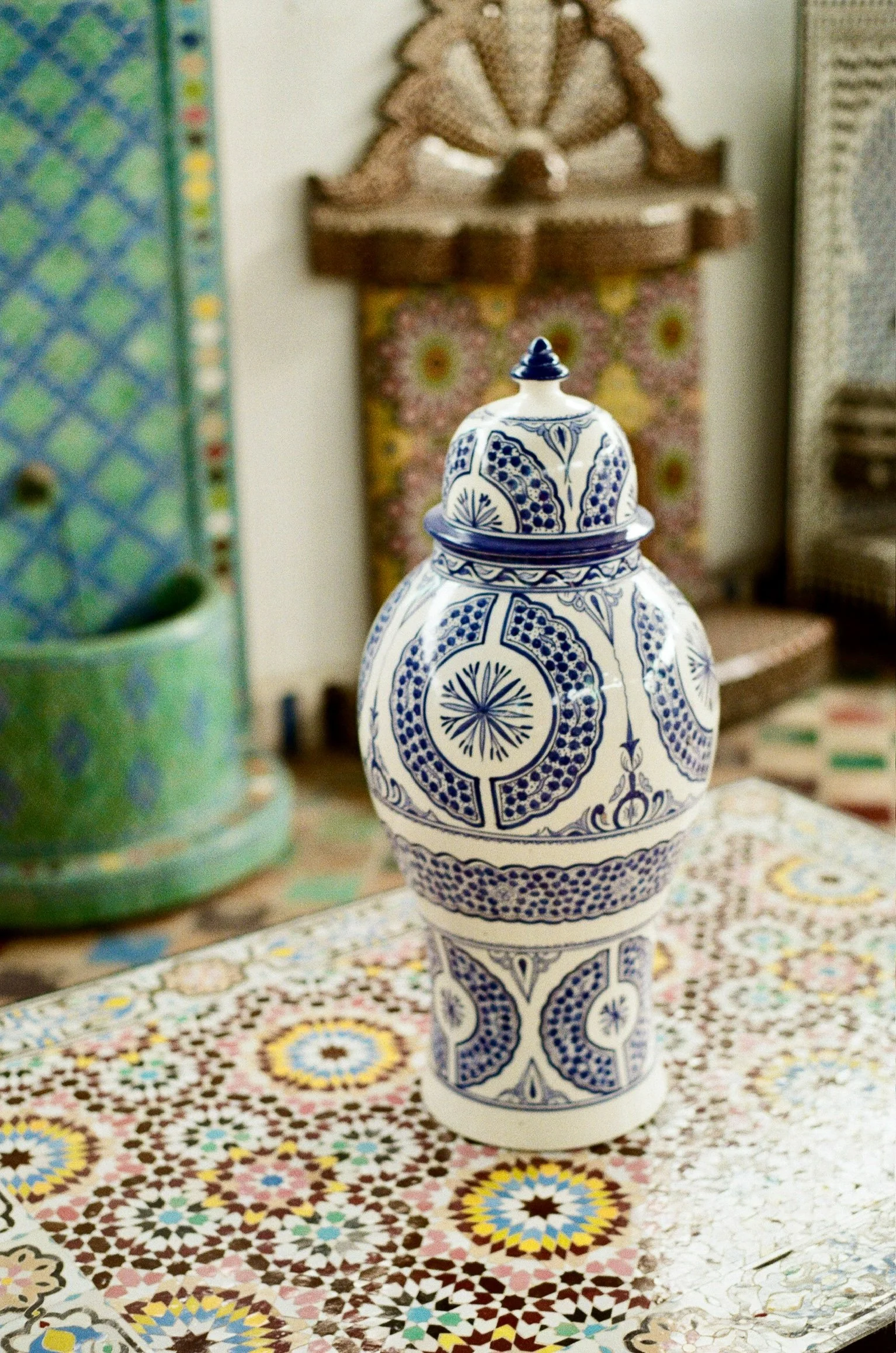 A decorative white and blue ceramic vase with intricate patterns, placed on a colorful patterned tablecloth, with various artisanal objects and artwork in the background.