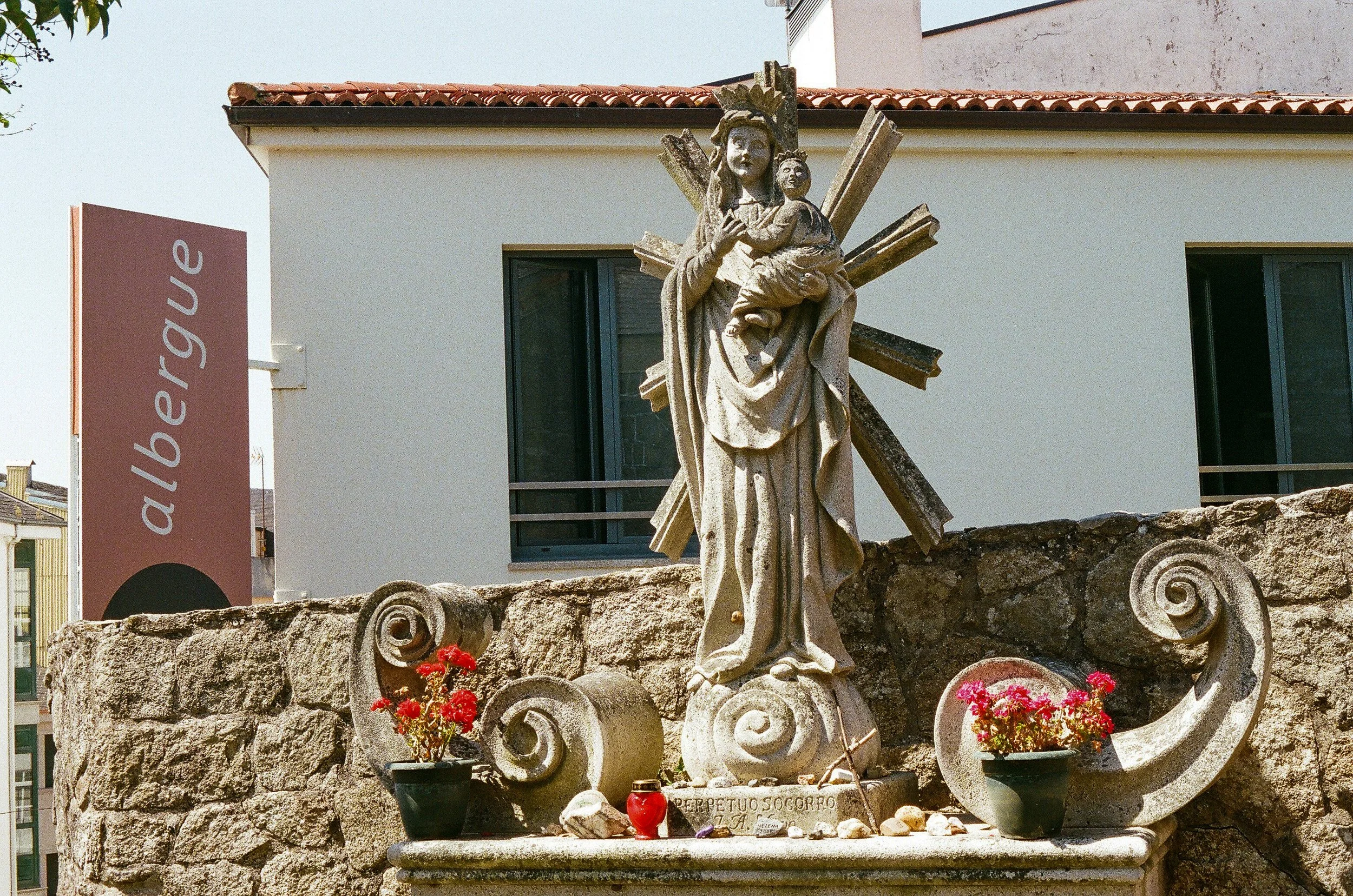 Statue of a woman holding a child, with star-like rays behind her, on a stone base surrounded by red flowers and various small items, in front of a modern building with a sign that reads 'auliergue' or similar.