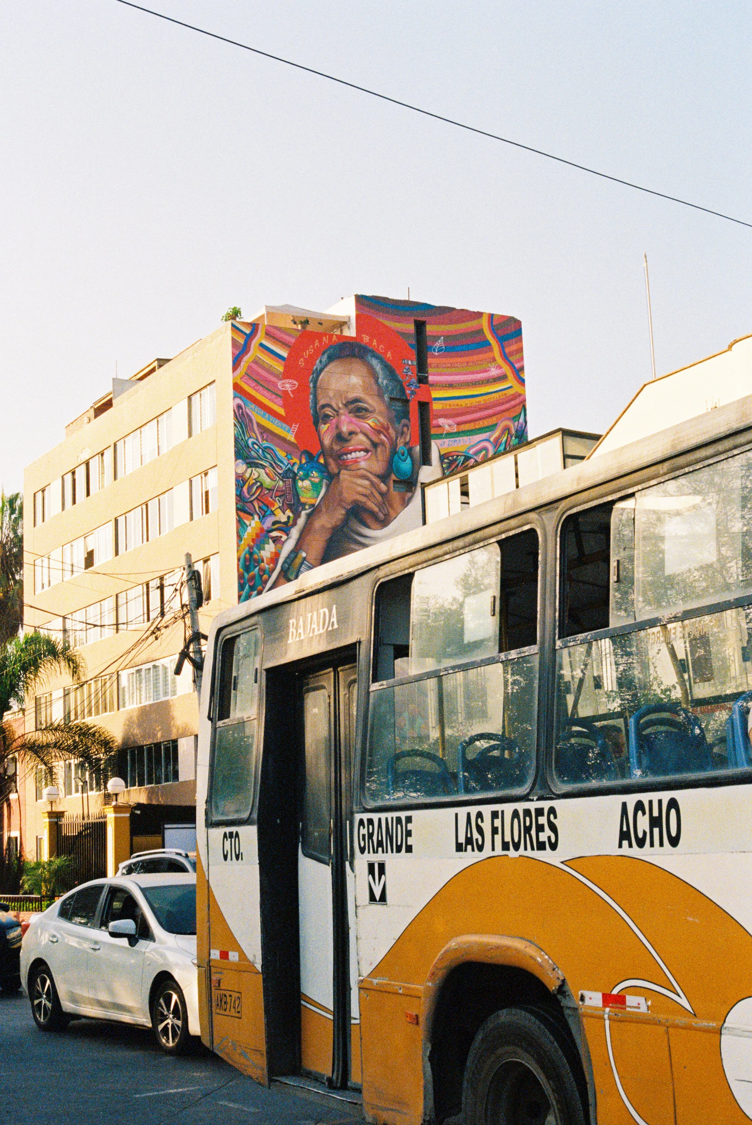 A colorful mural of a woman's face is painted on the side of a bus, with the city buildings and a white car in the background.