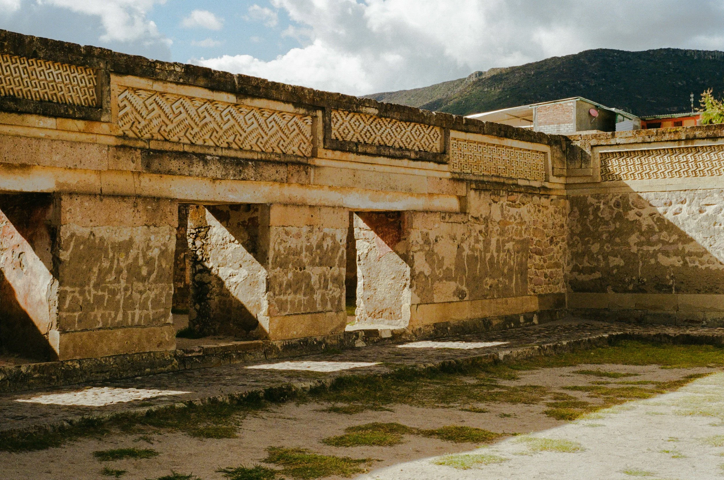 Ancient stone structure with intricate geometric carvings on the upper part of the walls, set against a backdrop of mountains and a cloudy sky.