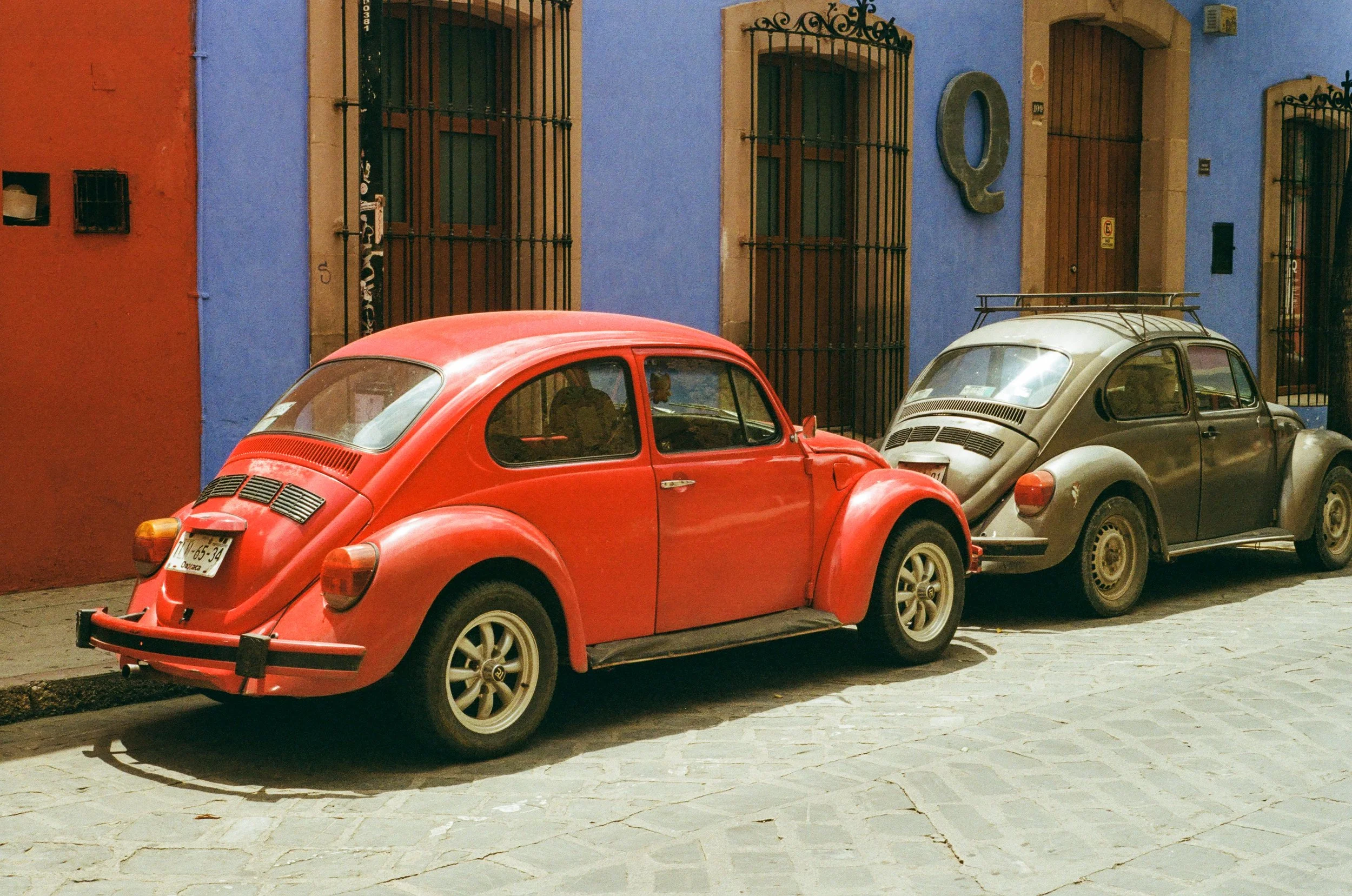 Two vintage Volkswagen Beetle cars parked on a cobblestone street in front of colorful buildings painted in red, purple, and blue. The buildings have barred windows and a one large decorative letter 'Q' on the wall.