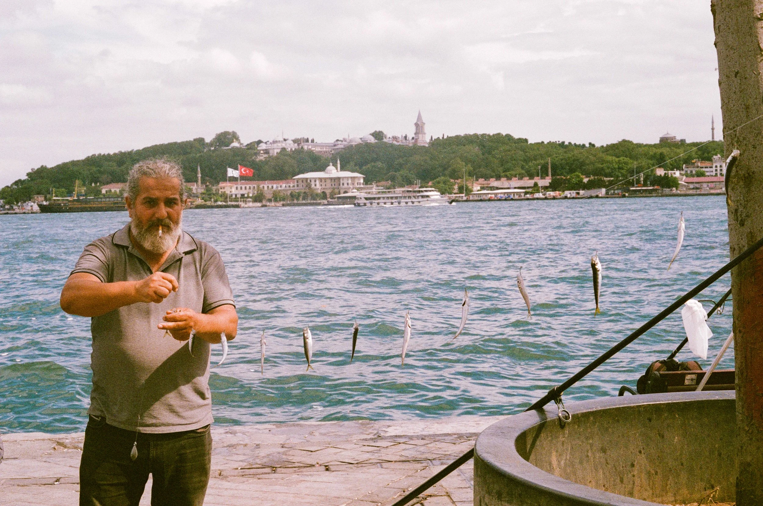 A man with gray hair and beard fishing by the water, hanging a string of small fish that he caught, with a cityscape and watercraft in the background.