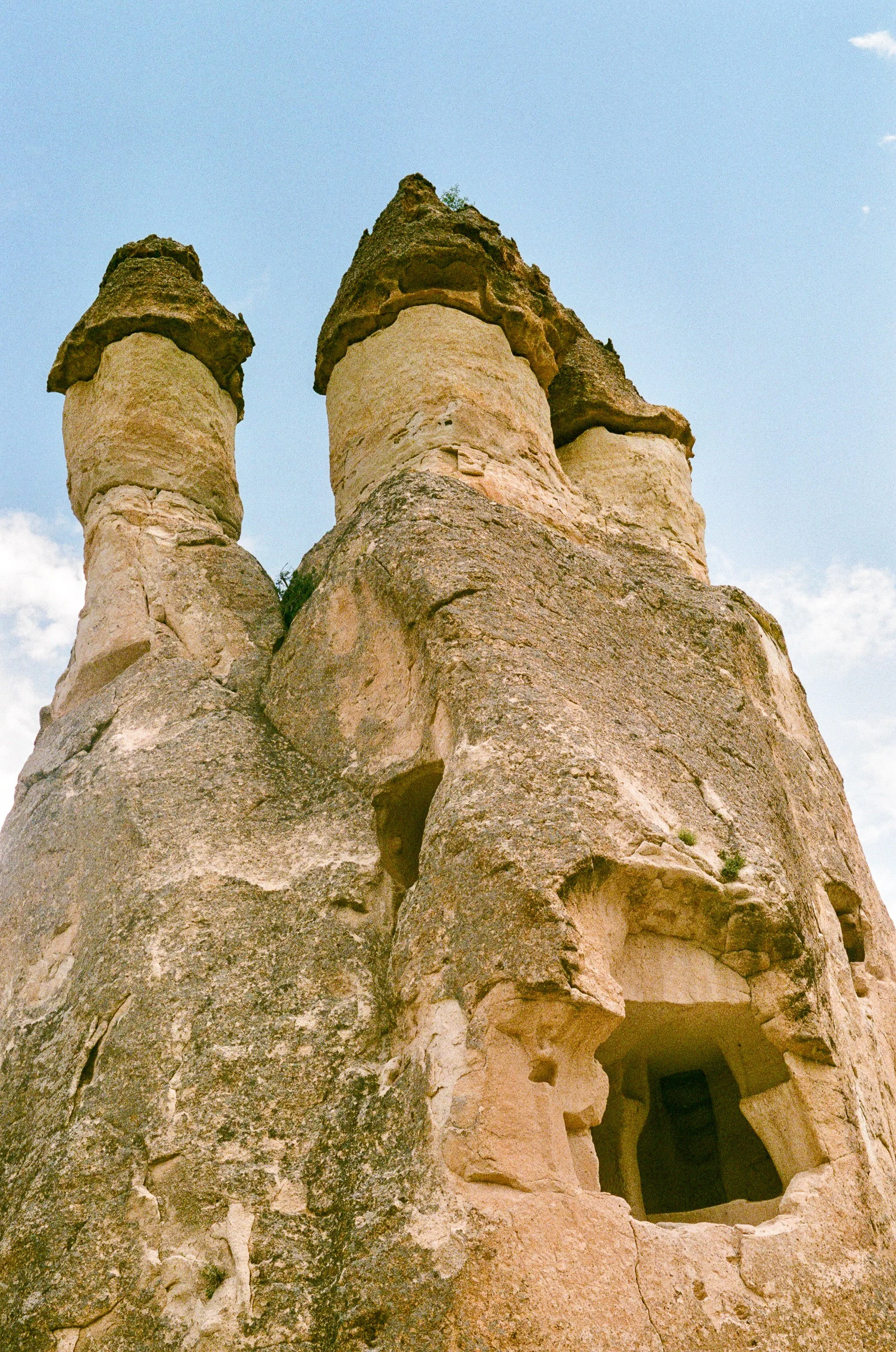 A tall rock formation with multiple pointed peaks against a partly cloudy sky, with a cave opening near the bottom right.