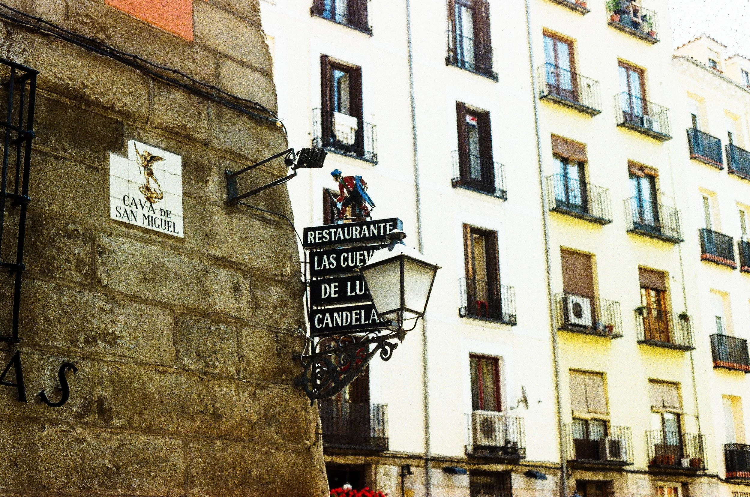 Signage on a building in an urban setting, including a sign for a restaurant called 'Las Cueva de L...' and a sign for 'Cava de San Miguel'. The background shows a multi-story residential building with balcony windows and flower boxes.