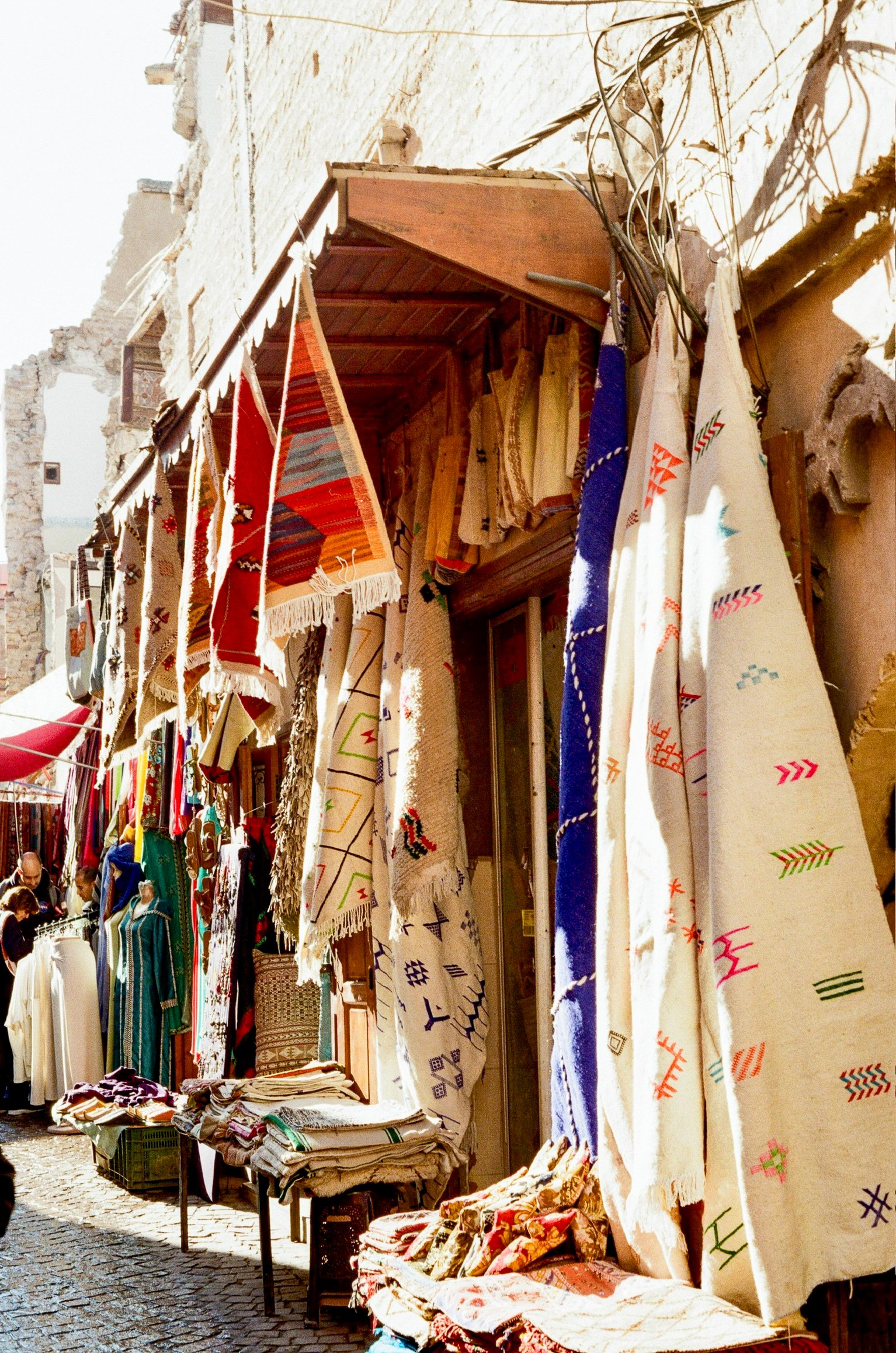 Outdoor market stall displaying colorful woven and embroidered textiles hanging on a wall, with tables below holding folded fabrics, on a cobblestone street.