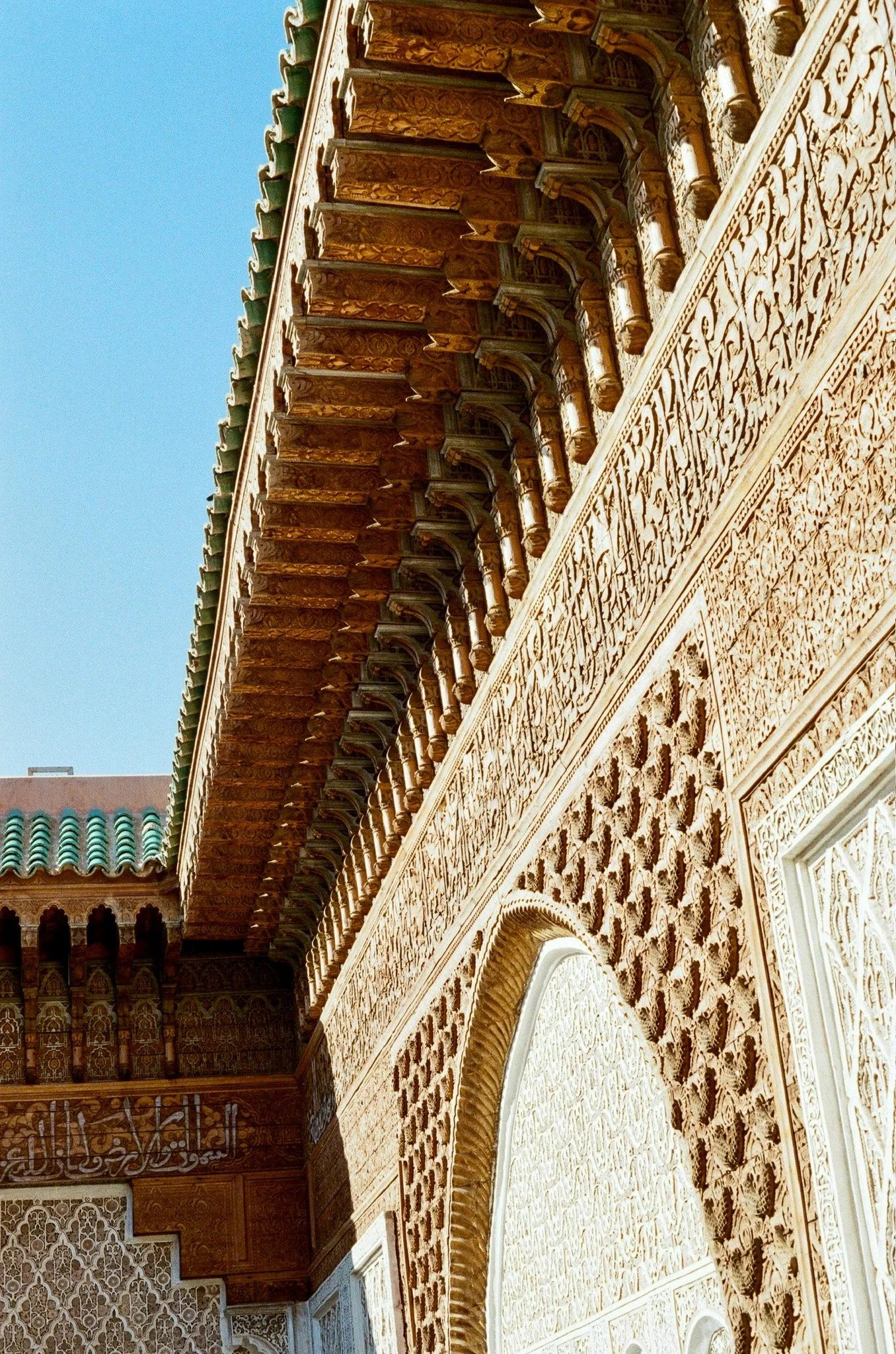 Close-up view of ornate architectural details on a building, including intricate carvings, arches, columns, and decorative patterns, with a portion of the roof visible against a clear blue sky.