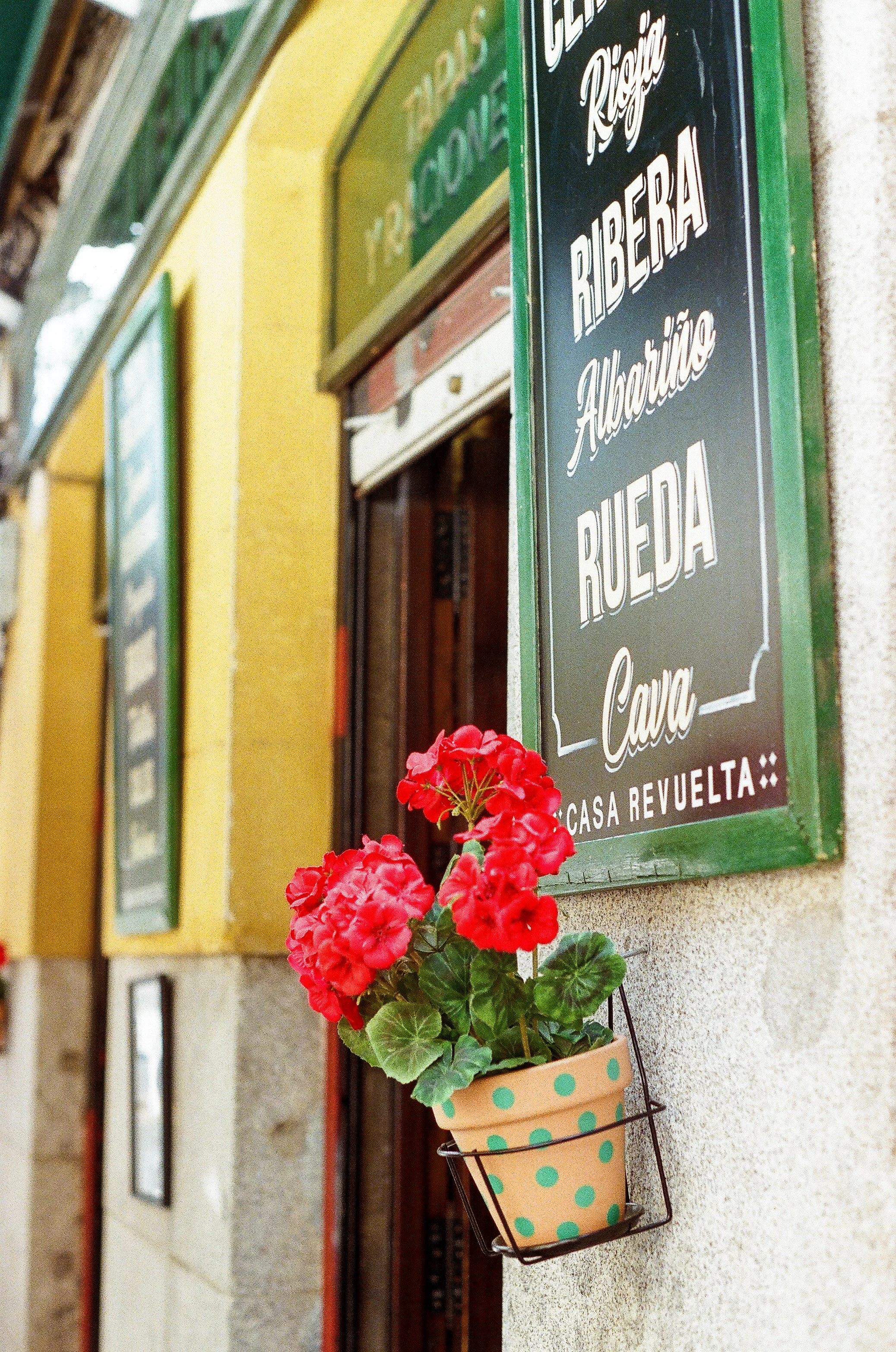 A decorative sign on a yellow wall advertising a restaurant or bar called Casa Revuelta, with a small potted plant of red flowers attached to the wall below the sign. The sign mentions 'Ribera' and 'Albaicín' among other words.