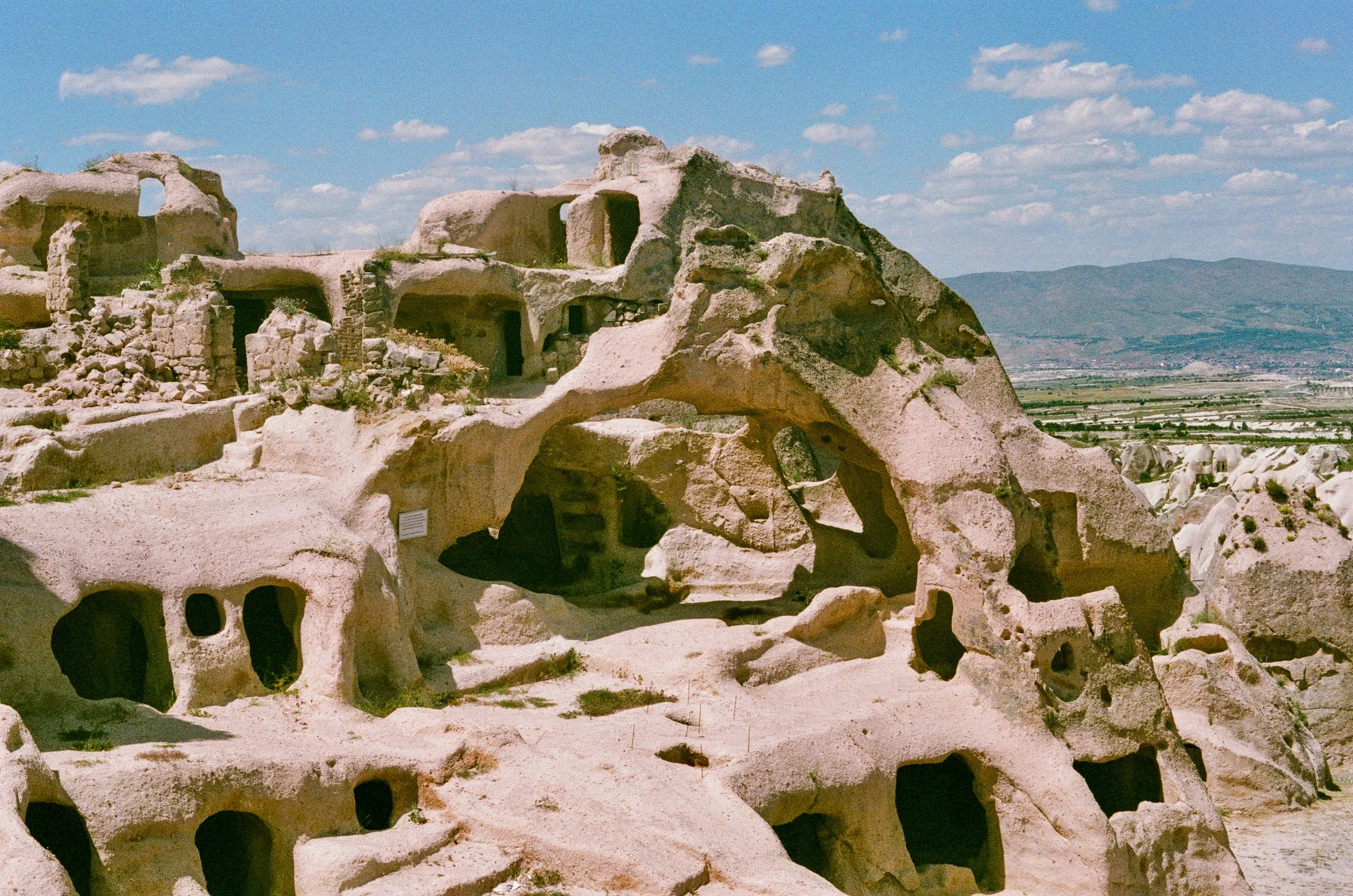 Ancient rock formations and cave dwellings in a desert landscape with a mountain range in the distance.