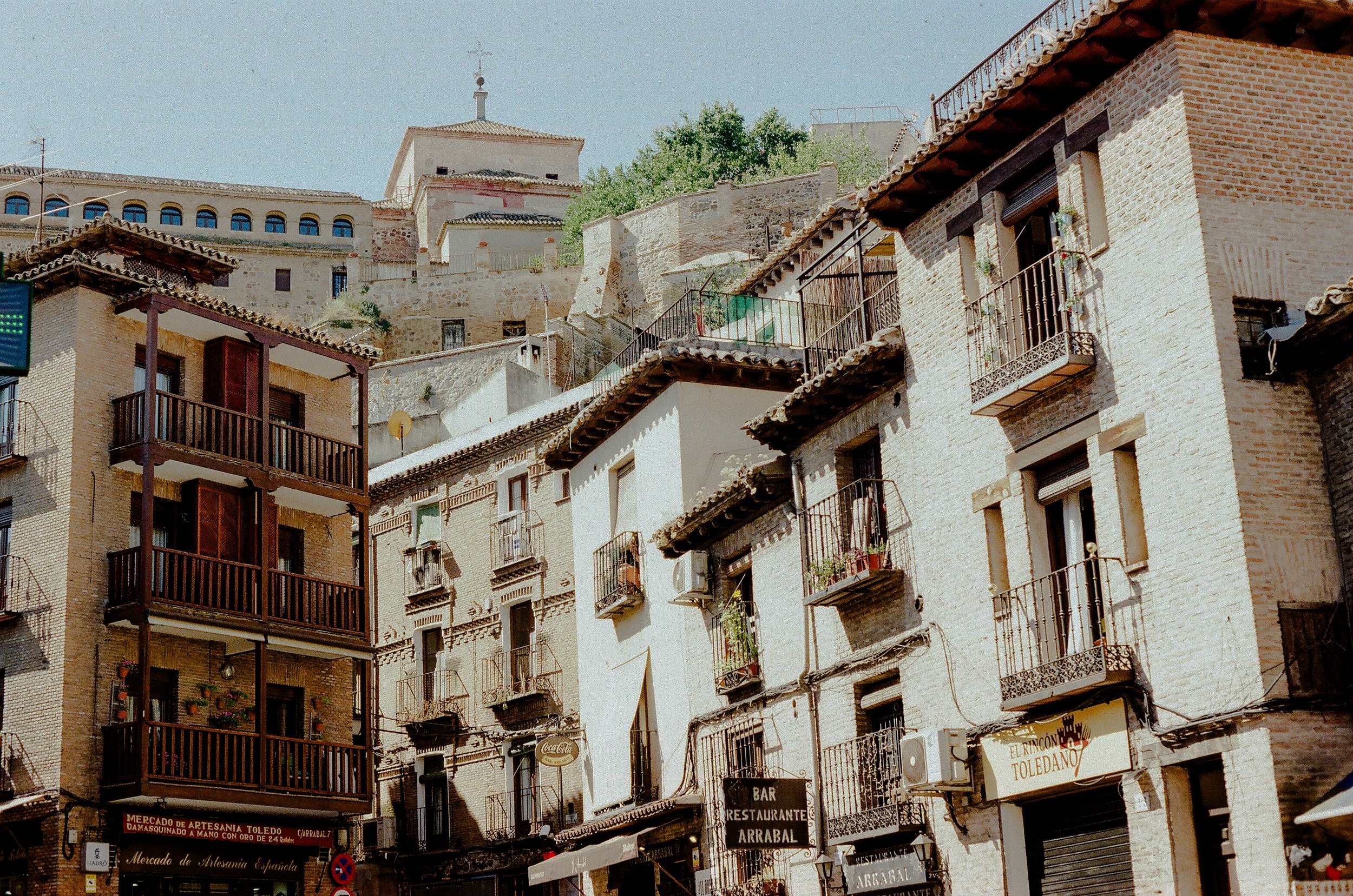 A view of multi-story buildings with balconies and signs at street level in Toledo, Spain, with a clear sky and some greenery visible in the background.