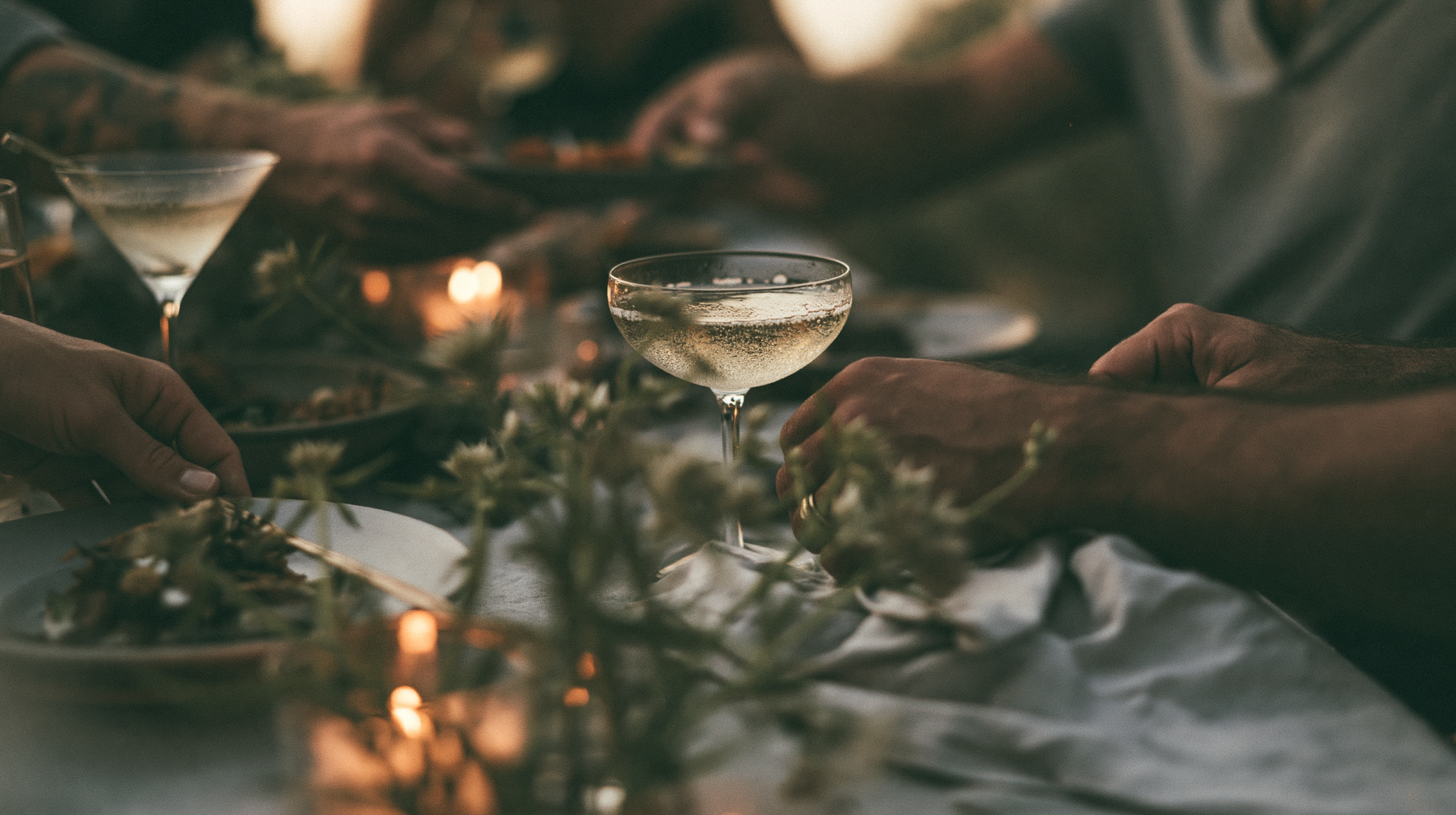 A close-up of a table with a cocktail glass and hands reaching for food, with warm, ambient lighting and floral decorations.