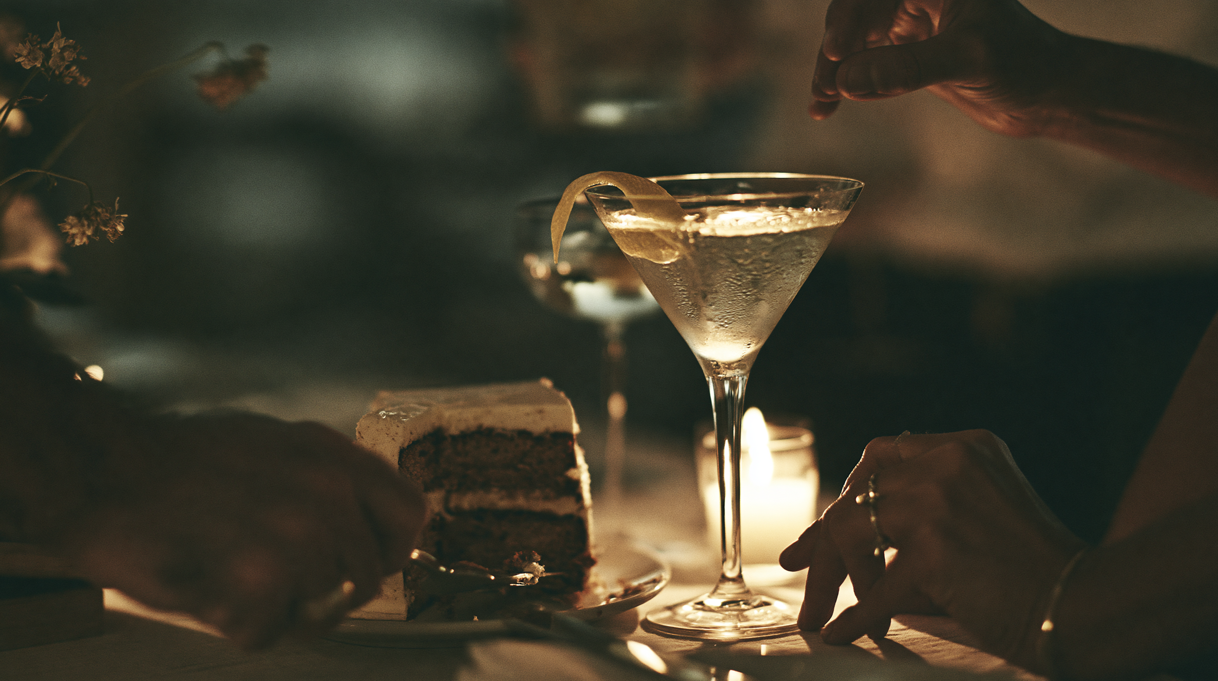 A person preparing a cocktail at a dimly lit party, with a slice of lemon on the rim of a martini glass, a layered cake, and a candle on the table.